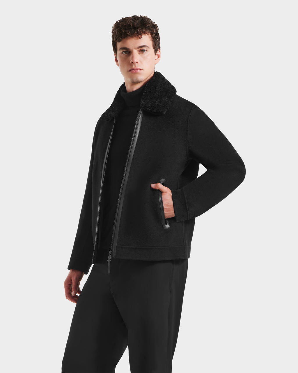 A young man with short curly hair models the LINCOLN - 6125038 Black coat by Rudsak, featuring a removable faux fur collar, layered over a black sweater and pants against a plain white background.