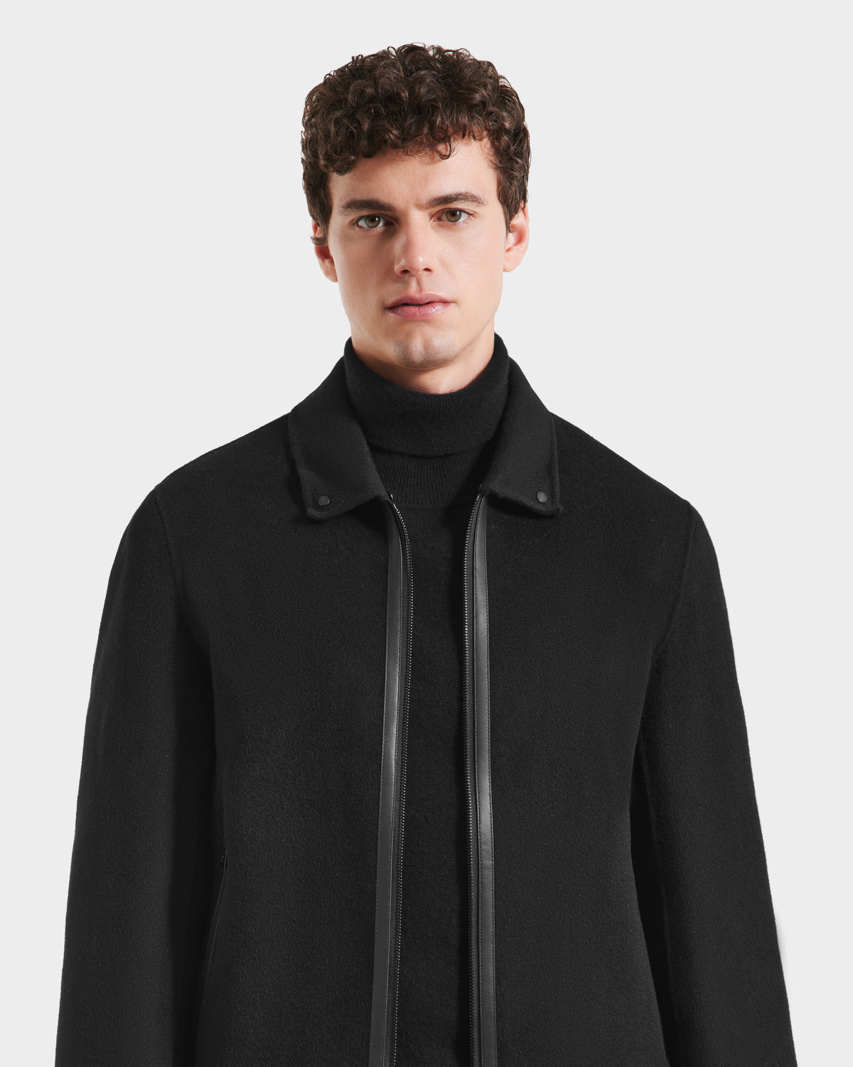 A young man with curly brown hair models the Rudsak LINCOLN - 6125038 Black coat, featuring a black turtleneck and a black wool design with leather trim, standing against a plain white background.