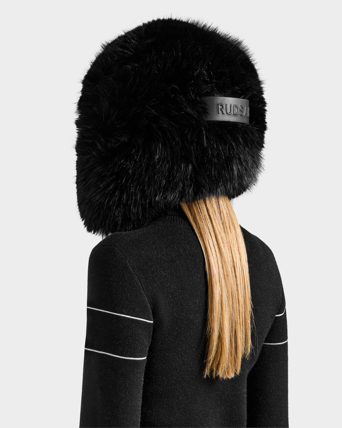 A person with long blonde hair wears the Rudsak LIRA - 8425024 Black faux fur aviator hat with "RUSSI" on the back, paired with a black sweater with white sleeve stripes, shown from behind for an effortless après-ski style.