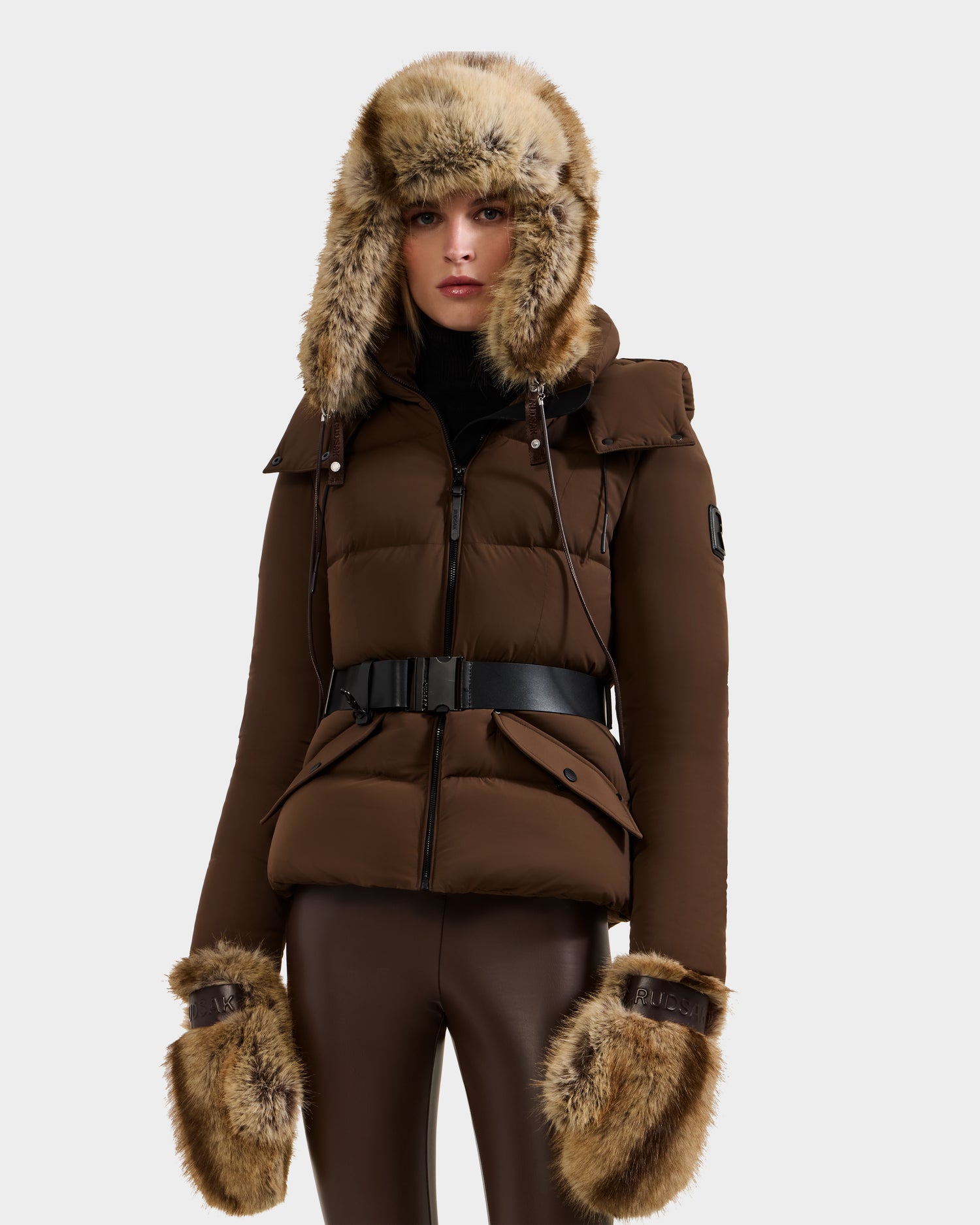 A person dressed in Rudsak's LIRA - 8425024 Mix Mocha puffer jacket with a belted waist, matching pants, a fur-trimmed hood, après-ski style fur mittens, and a faux fur aviator hat stands against a plain white background.
