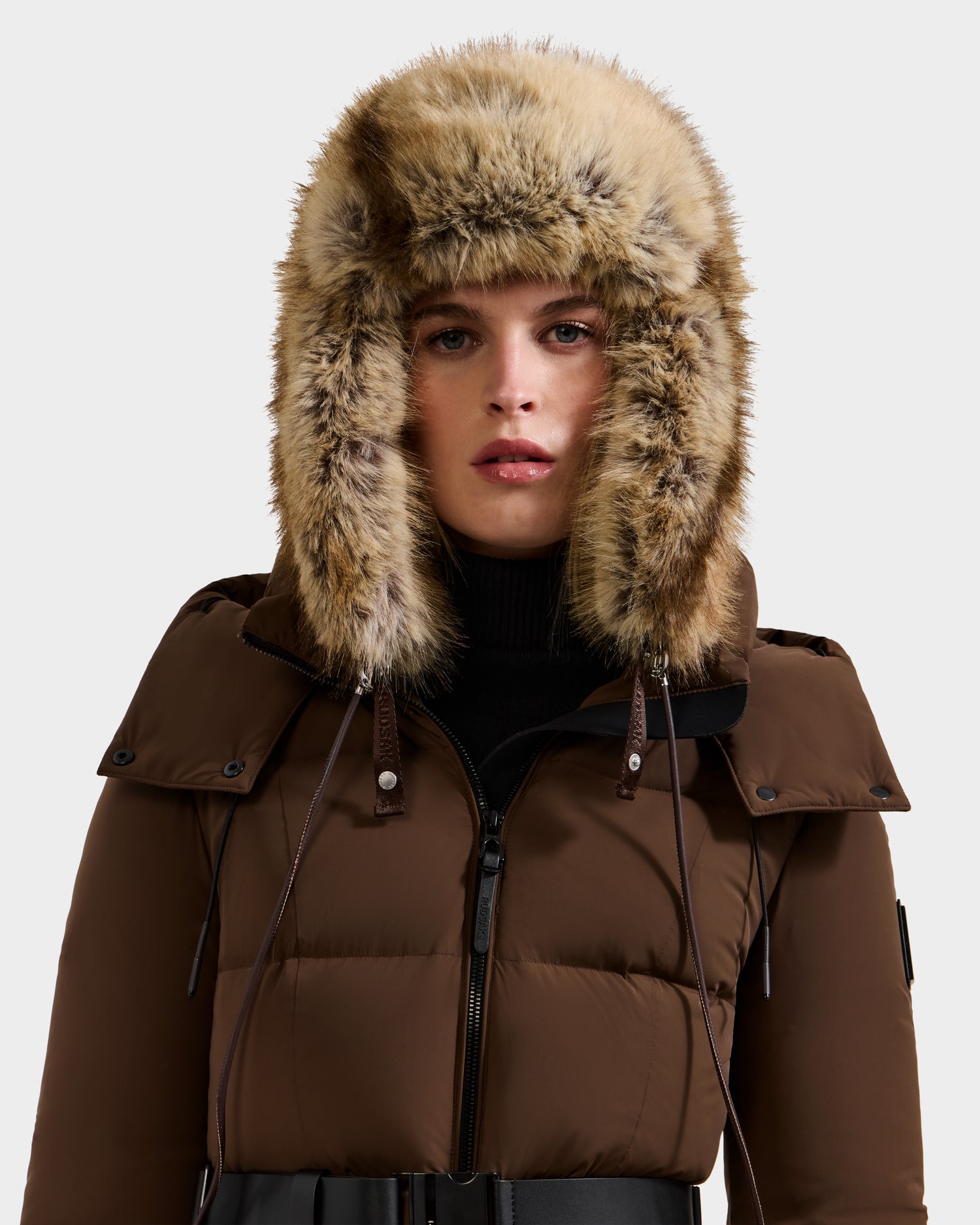 A woman in the Rudsak LIRA - 8425024 Mix Mocha puffer jacket and a faux fur aviator hat with ear flaps gazes at the camera, exuding cozy winter style against a plain light background.