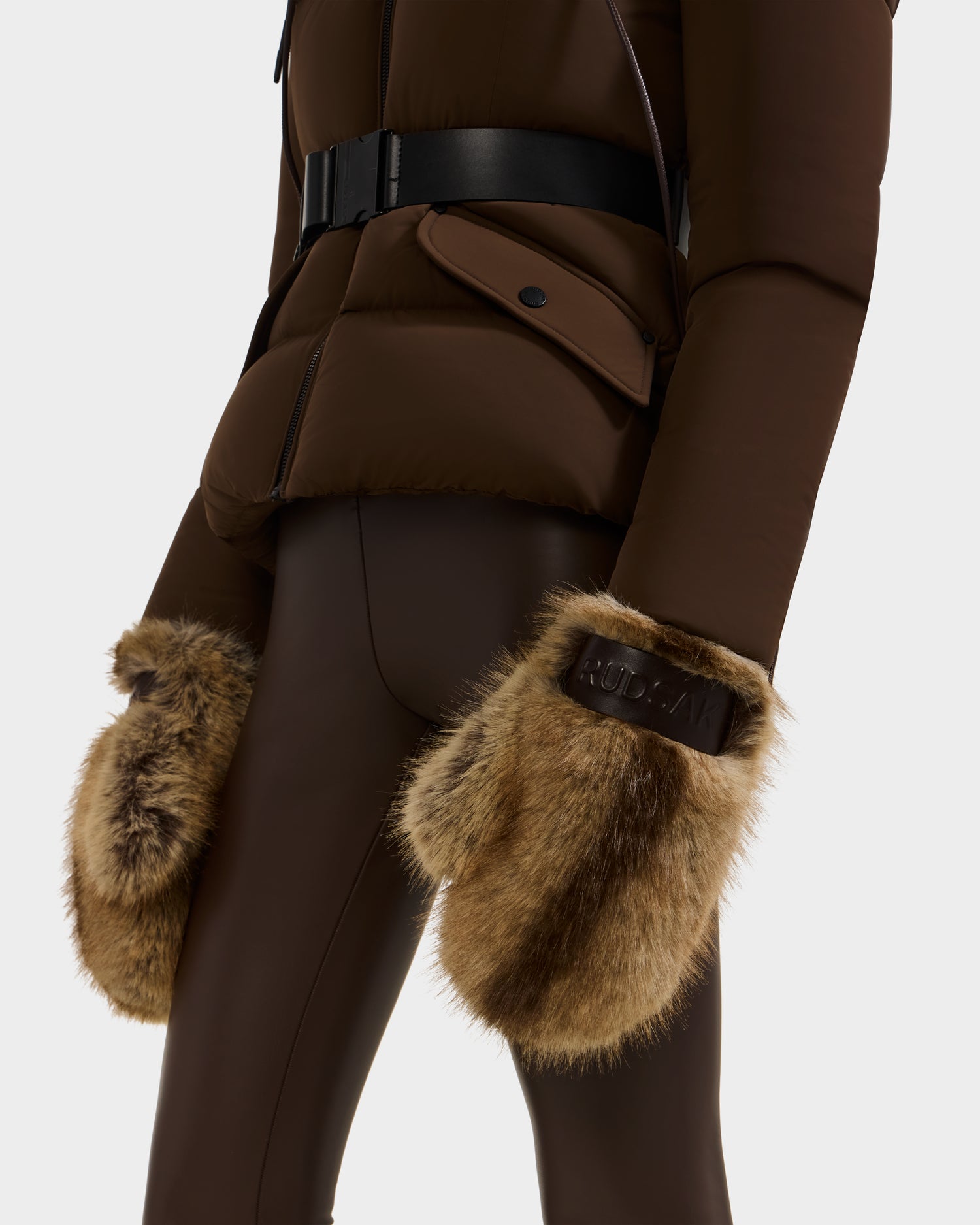 A person showcases winter style in the Rudsak LIRA - 8425024 Mix Mocha brown belted puffer jacket, fitted pants, and faux fur mittens with logo-embossed cuffs. The image is cropped at shoulder and mid-thigh.