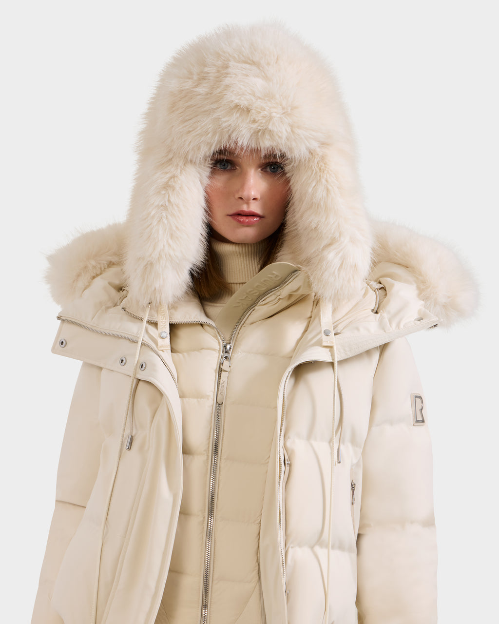 A woman models the Rudsak LIRA - 8425024 Vanilla, a cream puffer jacket with a fluffy fur hood, styled over a beige turtleneck and shown against a plain light background.