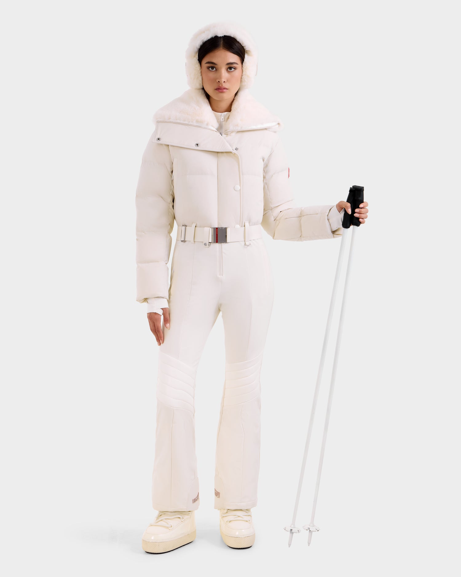 A woman models the LOVE - 8124724 Vanilla women's ski suit by Rudsak, featuring a fur-trimmed hood, belted jacket, and matching pants. She holds two ski poles and wears white boots against a plain light background.