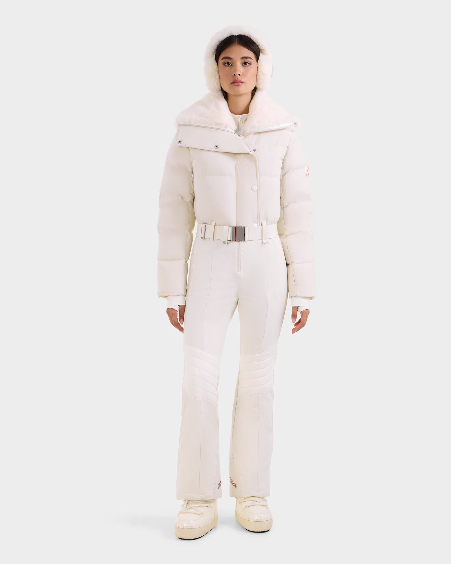 A woman faces forward in the Rudsak LOVE - 8124724 Vanilla, a stylish, fitted white one-piece ski suit with RECCO rescue tech, wide collar, matching hood, belted waist, and white boots against a plain light background.