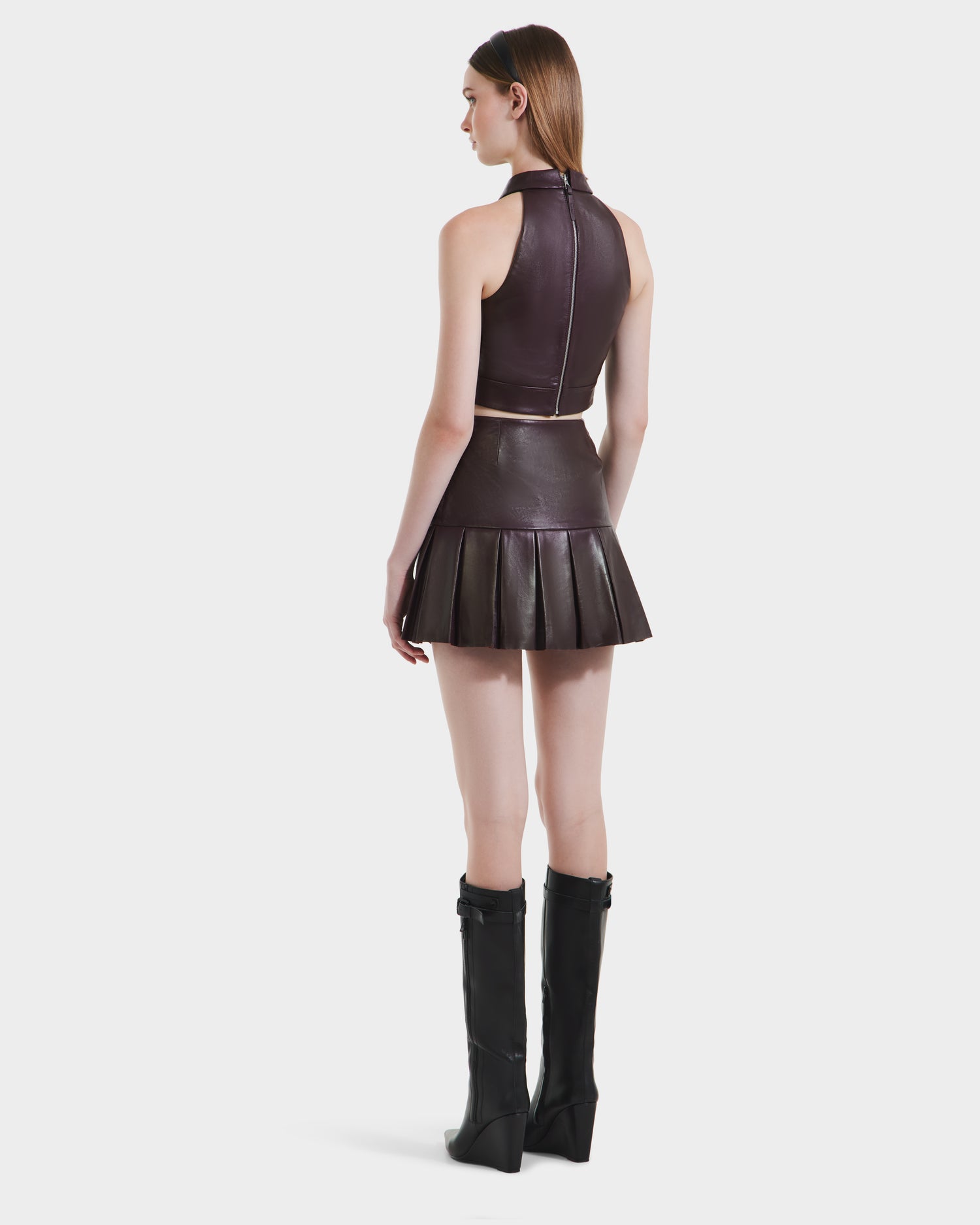 A woman faces away, wearing the Rudsak LOVELL - 8525557 Bordeaux premium leather sleeveless halter dress with a pleated skirt and black knee-high boots, set against a plain white background.