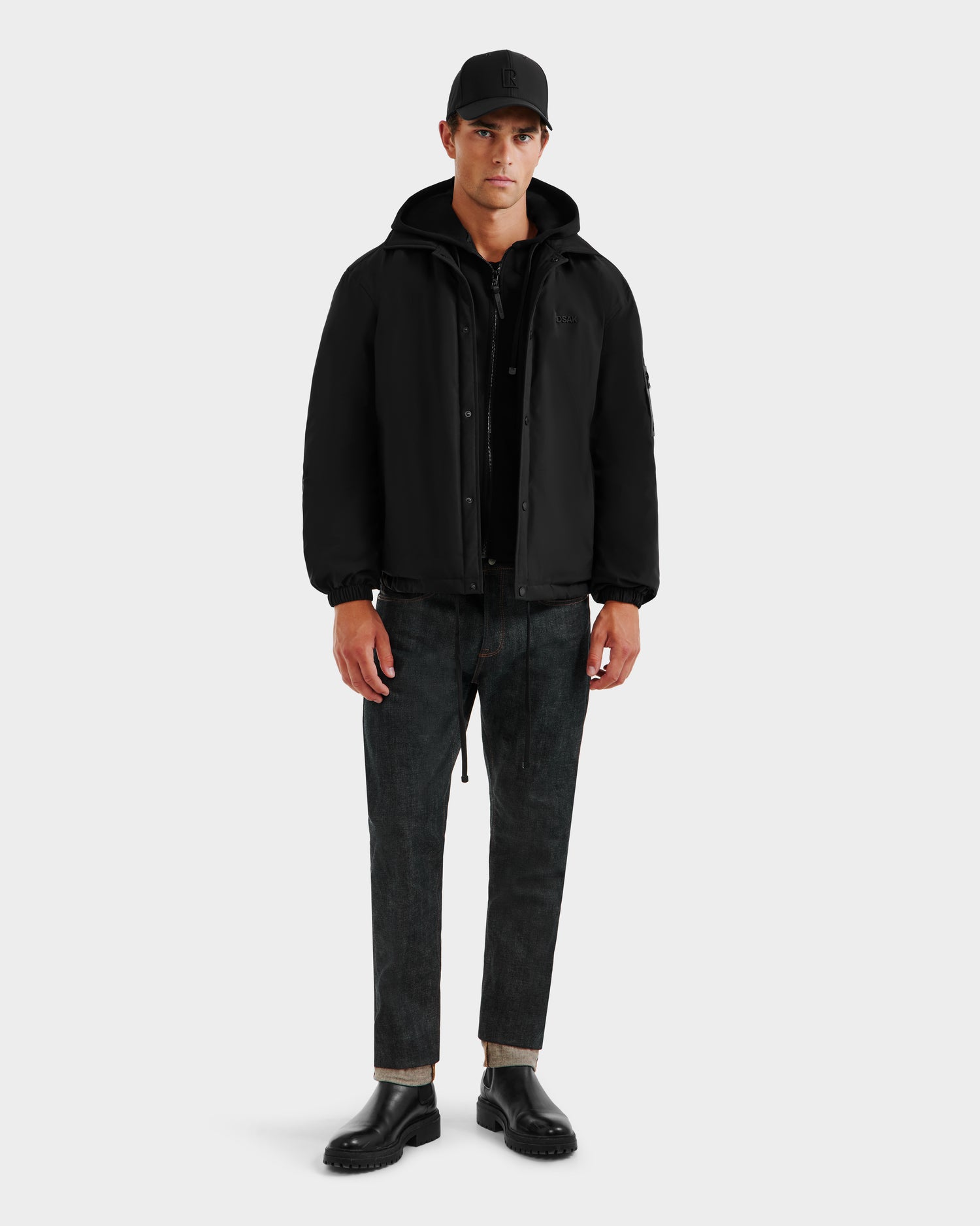 A person faces forward, wearing the Rudsak LUCA - 6125009 Black weather-resistant cap, a men’s hooded jacket, black shirt, cuffed dark jeans, and black shoes against a plain light gray background.