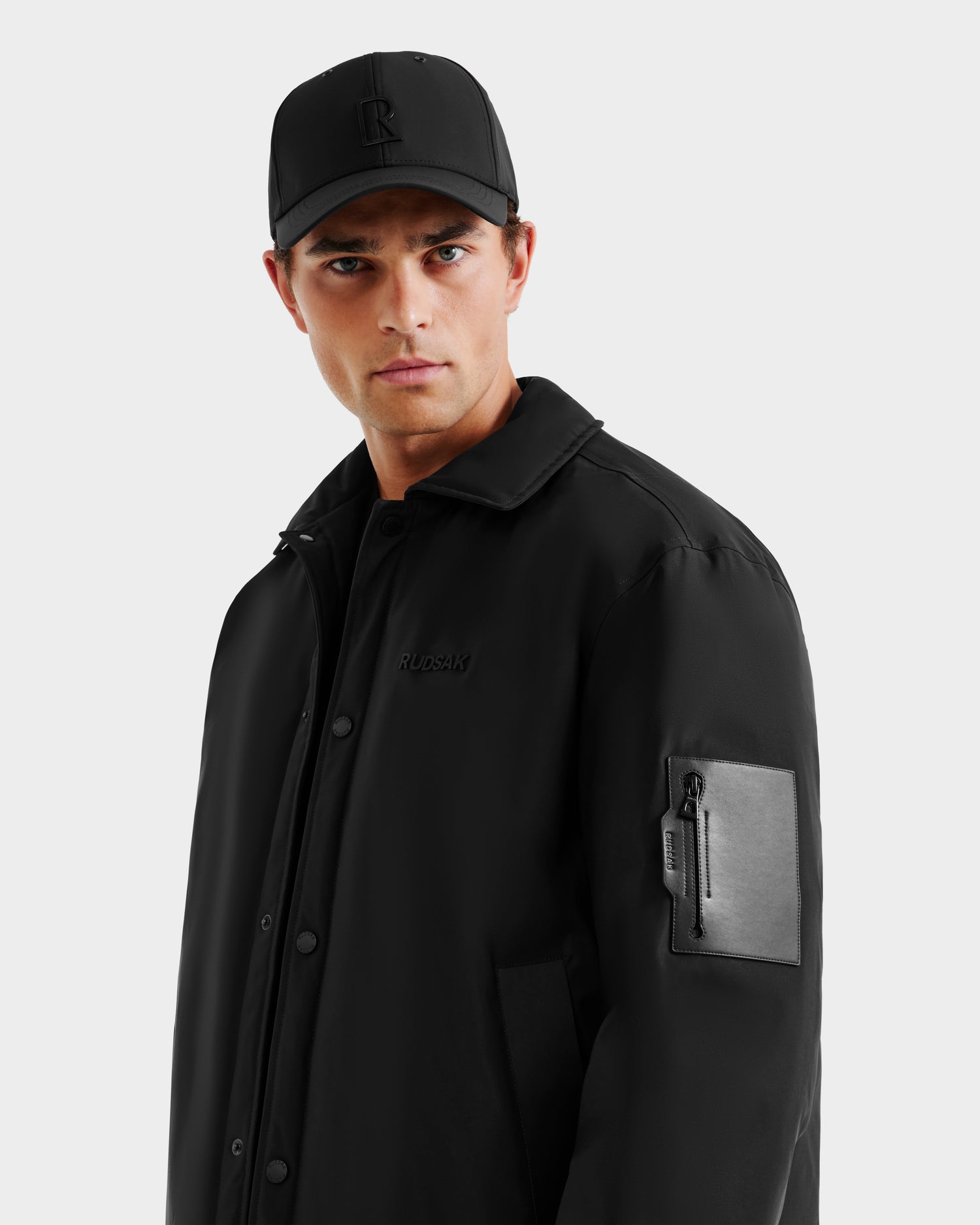 A man in the Rudsak LUCA - 6125009 Black weather-resistant men’s jacket with a zippered sleeve pocket stands against a light gray background, gazing seriously at the camera.