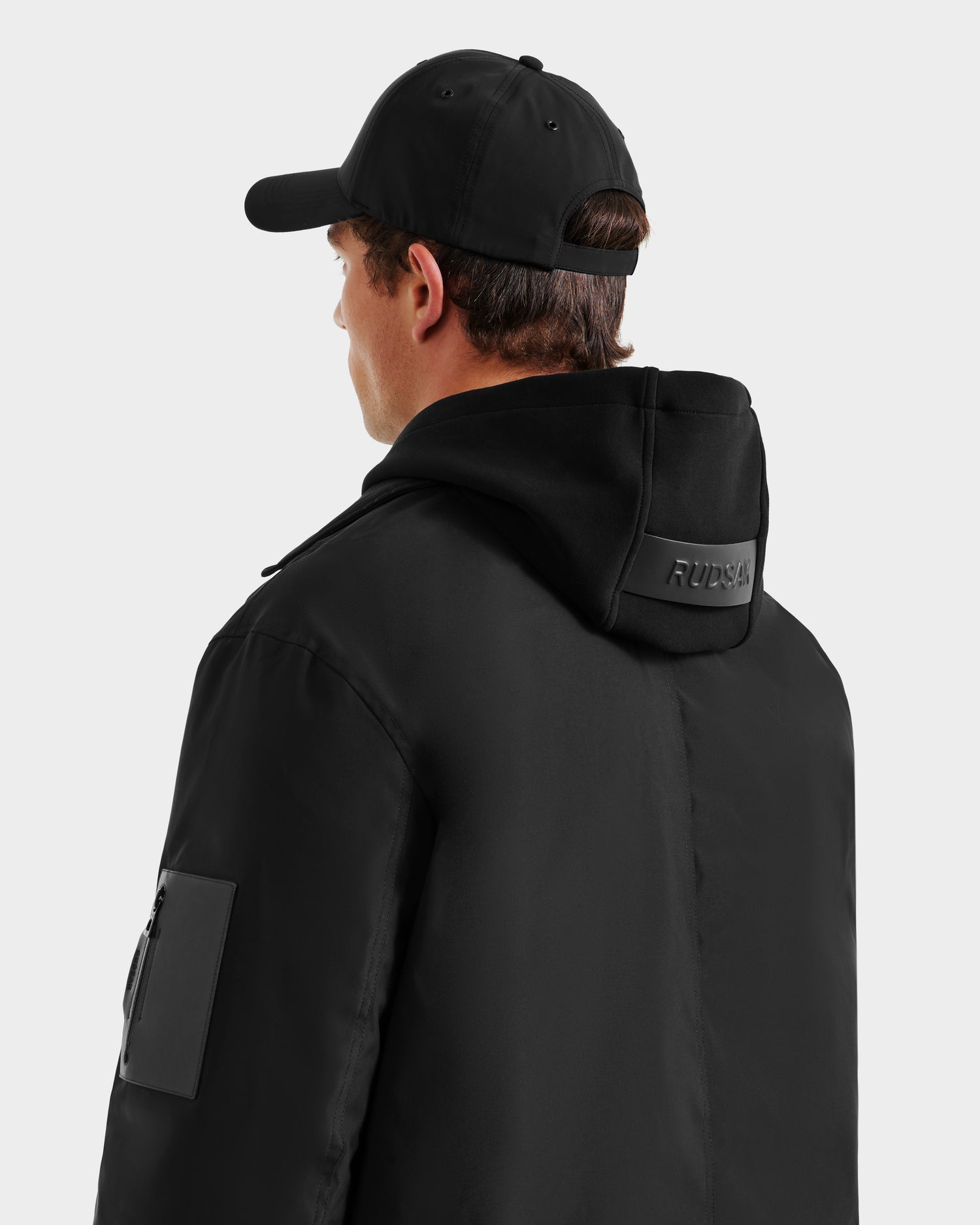 A person wearing the Rudsak LUCA - 6125009 weather-resistant black jacket with a hood and a black baseball cap, shown from the back on a plain light background.