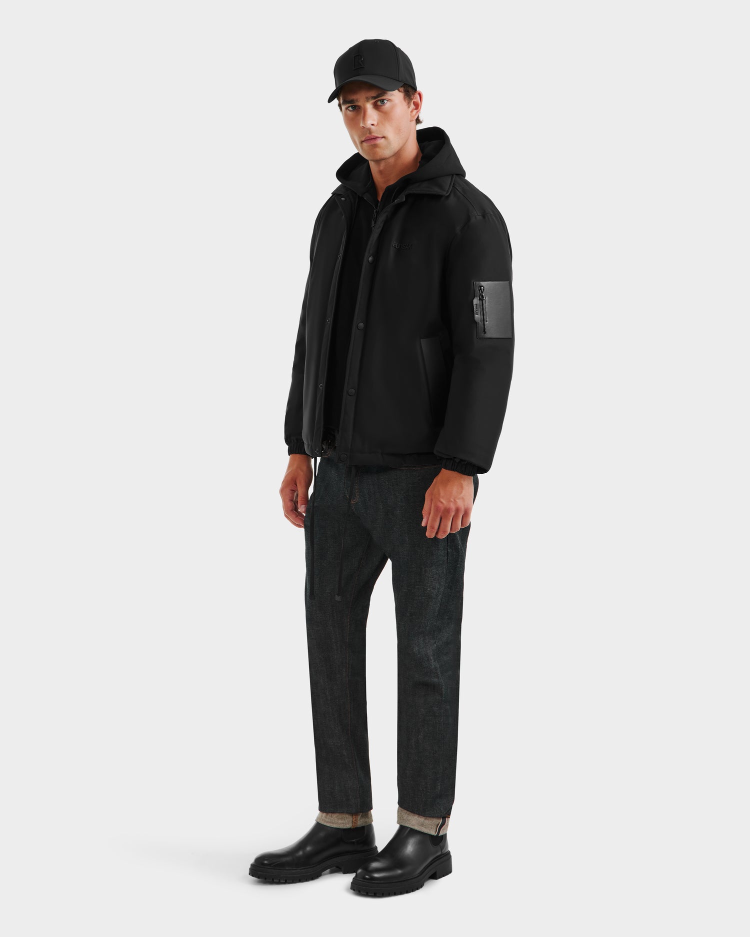 A man wearing the Rudsak LUCA - 6125009 Black weather-resistant insulated down mens jacket with a zippered sleeve pocket, paired with a black cap, dark cuffed jeans, and black boots stands against a plain white background.
