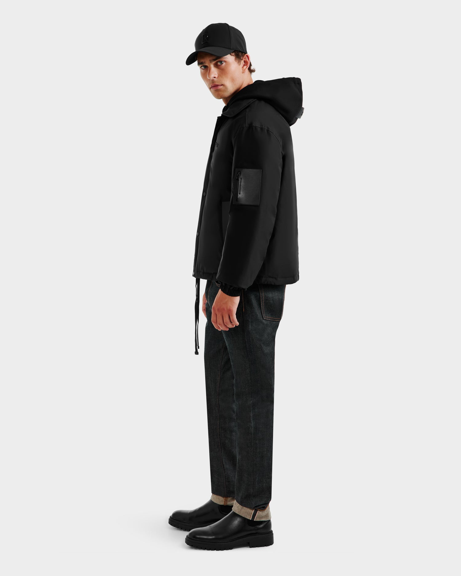 A man in the Rudsak LUCA - 6125009 Black weather-resistant men’s jacket, black cap, dark jeans, and black boots stands sideways against a plain white background.