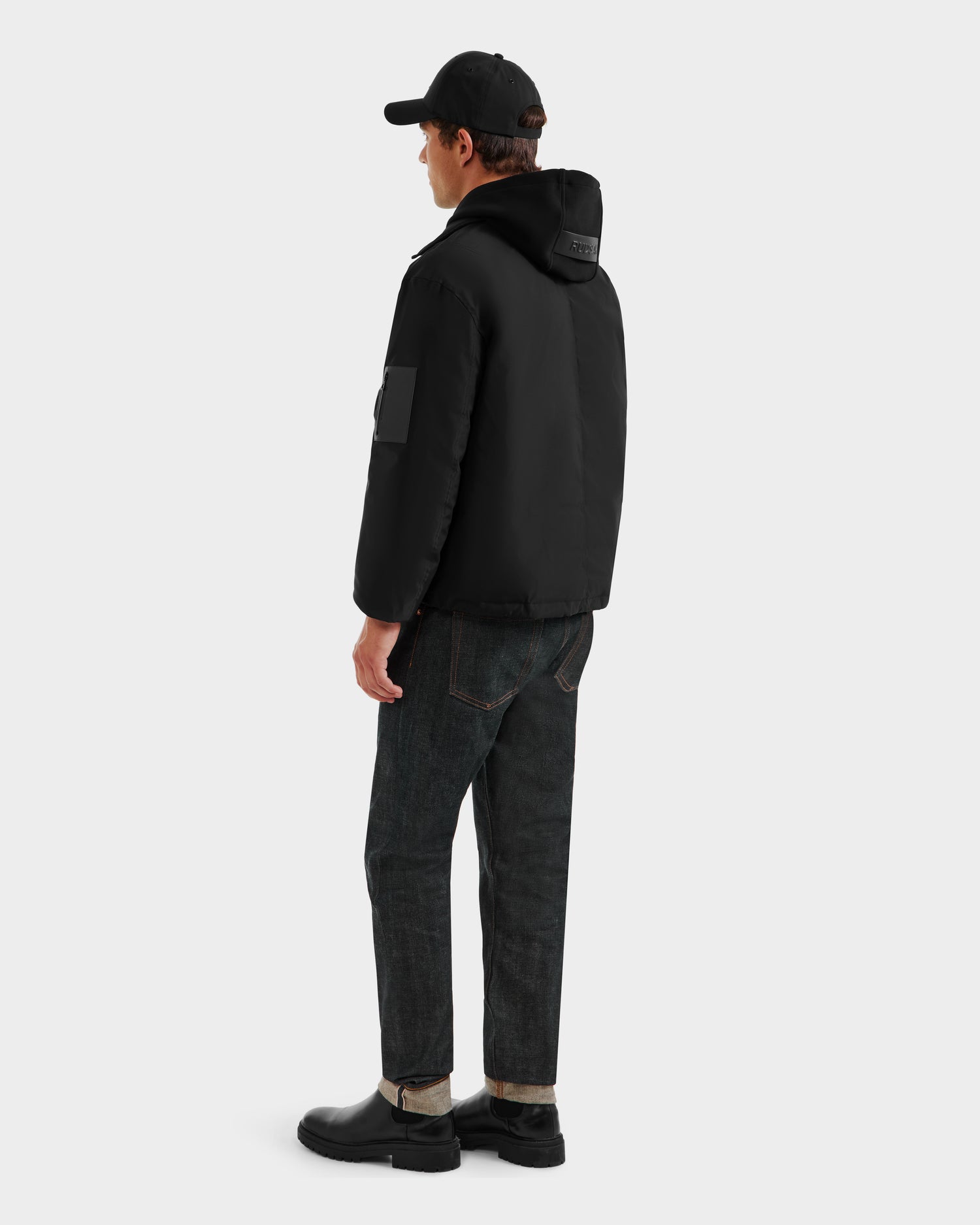 A person faces away, wearing the Rudsak LUCA - 6125009 black weather-resistant puffer jacket with a hood, paired with dark cuffed jeans, black boots, and a black baseball cap against a plain light background.