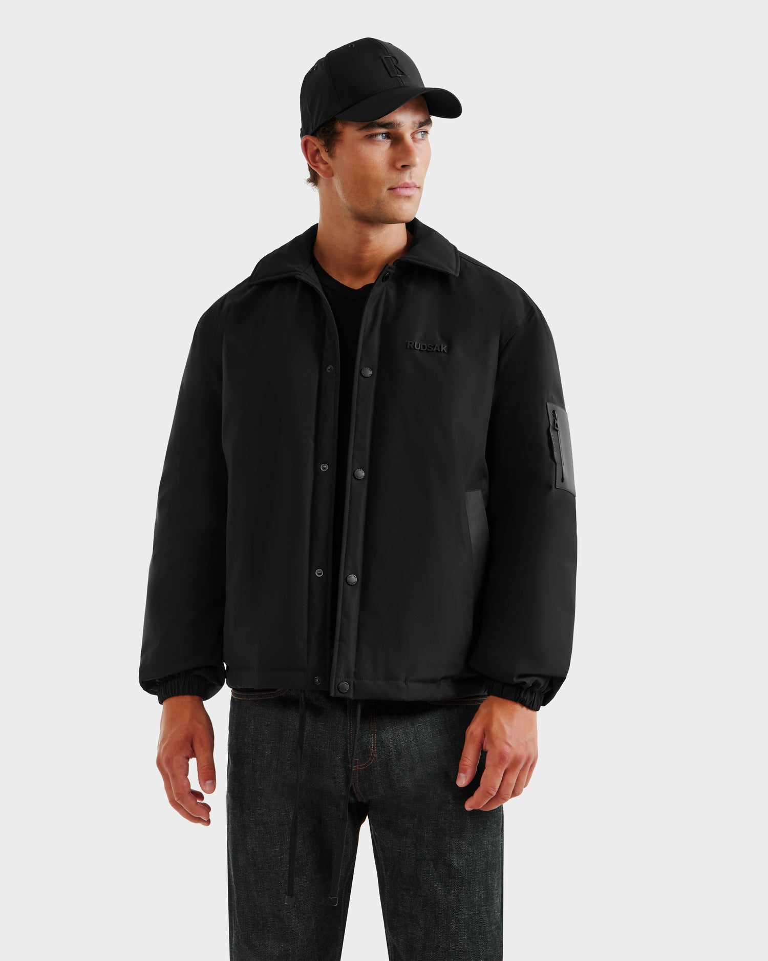 A man wearing the Rudsak LUCA - 6125009 Black weather-resistant men's jacket, black t-shirt, black baseball cap, and dark jeans stands against a plain light background, looking to the side with a neutral expression.