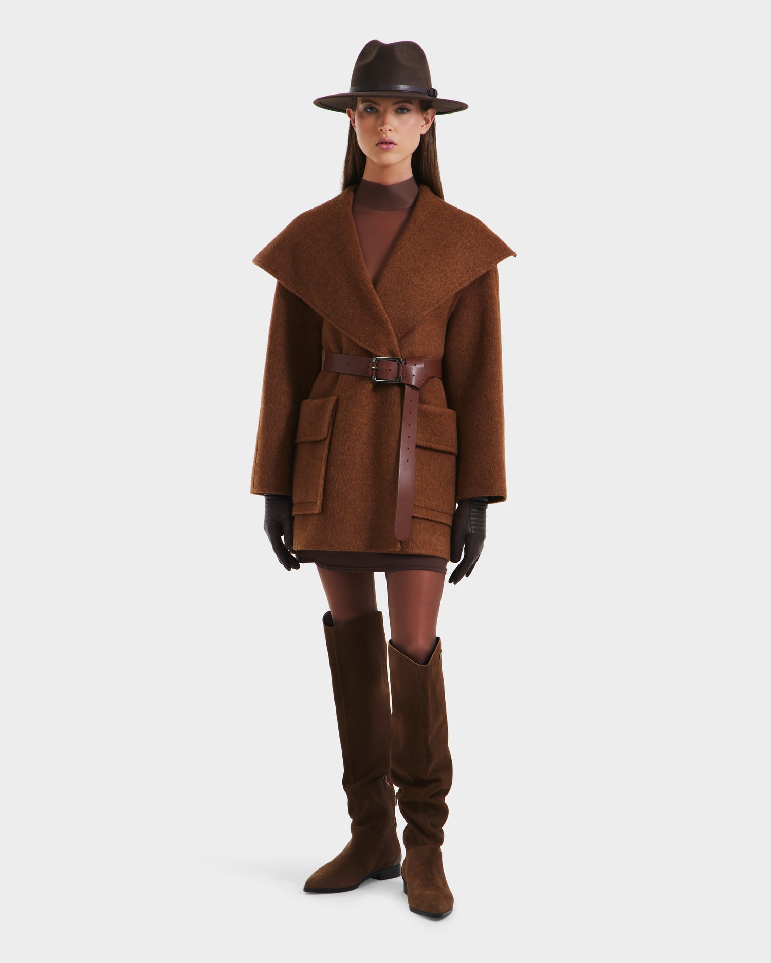 A woman wearing the Rudsak LUCIA - 8125633 Camel oversized wool jacket with large lapels and a vegan leather belt stands against a plain white background, styled with a wide-brimmed hat, brown gloves, mini dress, and knee-high boots.