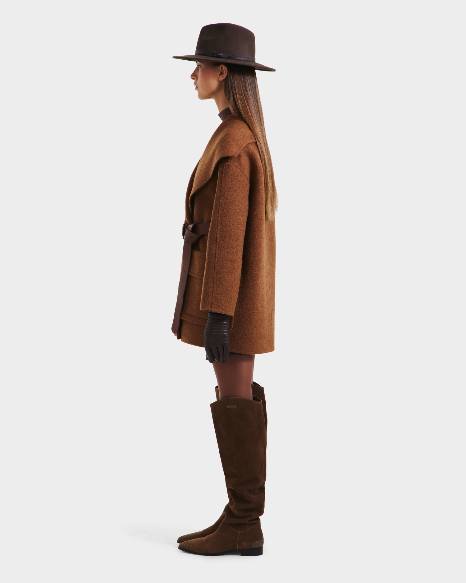 A woman stands in profile wearing the Rudsak LUCIA - 8125633 Camel wool jacket with a vegan leather belt, paired with brown gloves, knee-high boots, and tights, against a plain white background.