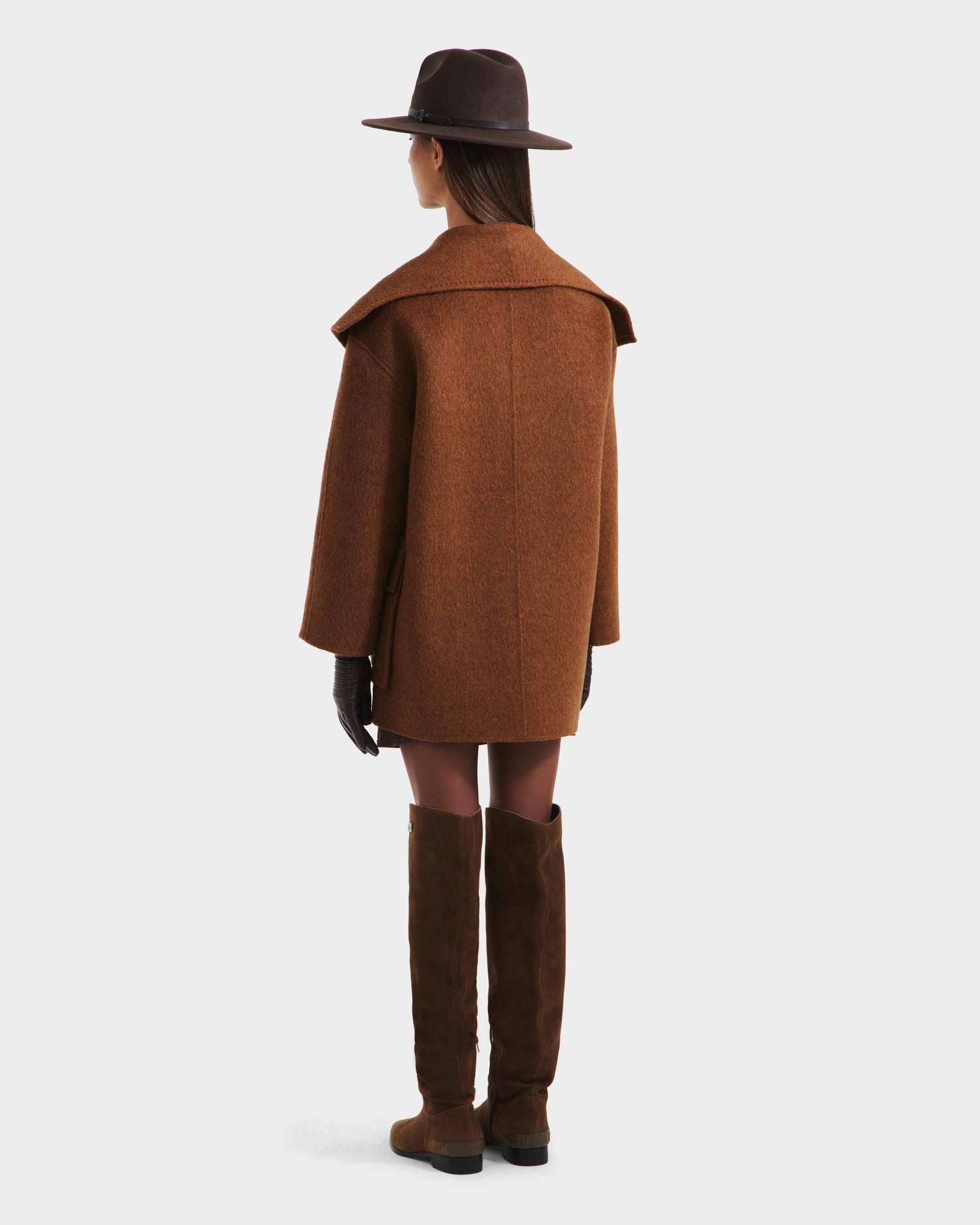 A woman faces away, wearing the Rudsak LUCIA - 8125633 Camel wool jacket as an oversized coat, paired with a brown wide-brimmed hat, black gloves, knee-high brown boots, and a vegan leather belt against a plain light background.