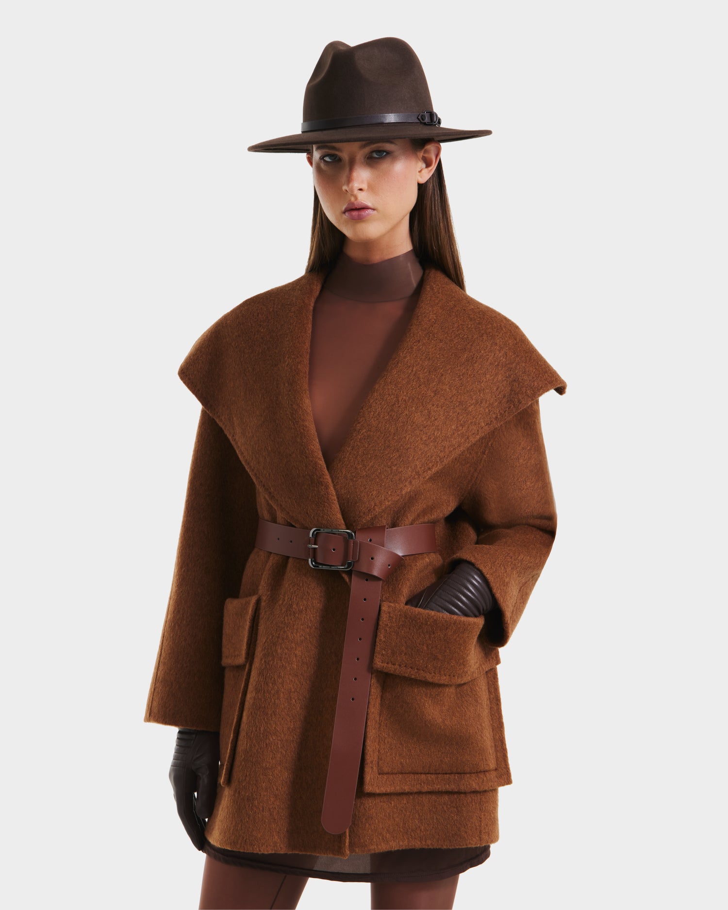 A woman stands against a plain white background wearing the Rudsak LUCIA - 8125633 Camel, an oversized women’s wool belted jacket with large pockets, styled with a wide-brimmed brown hat, brown gloves, and a brown dress.