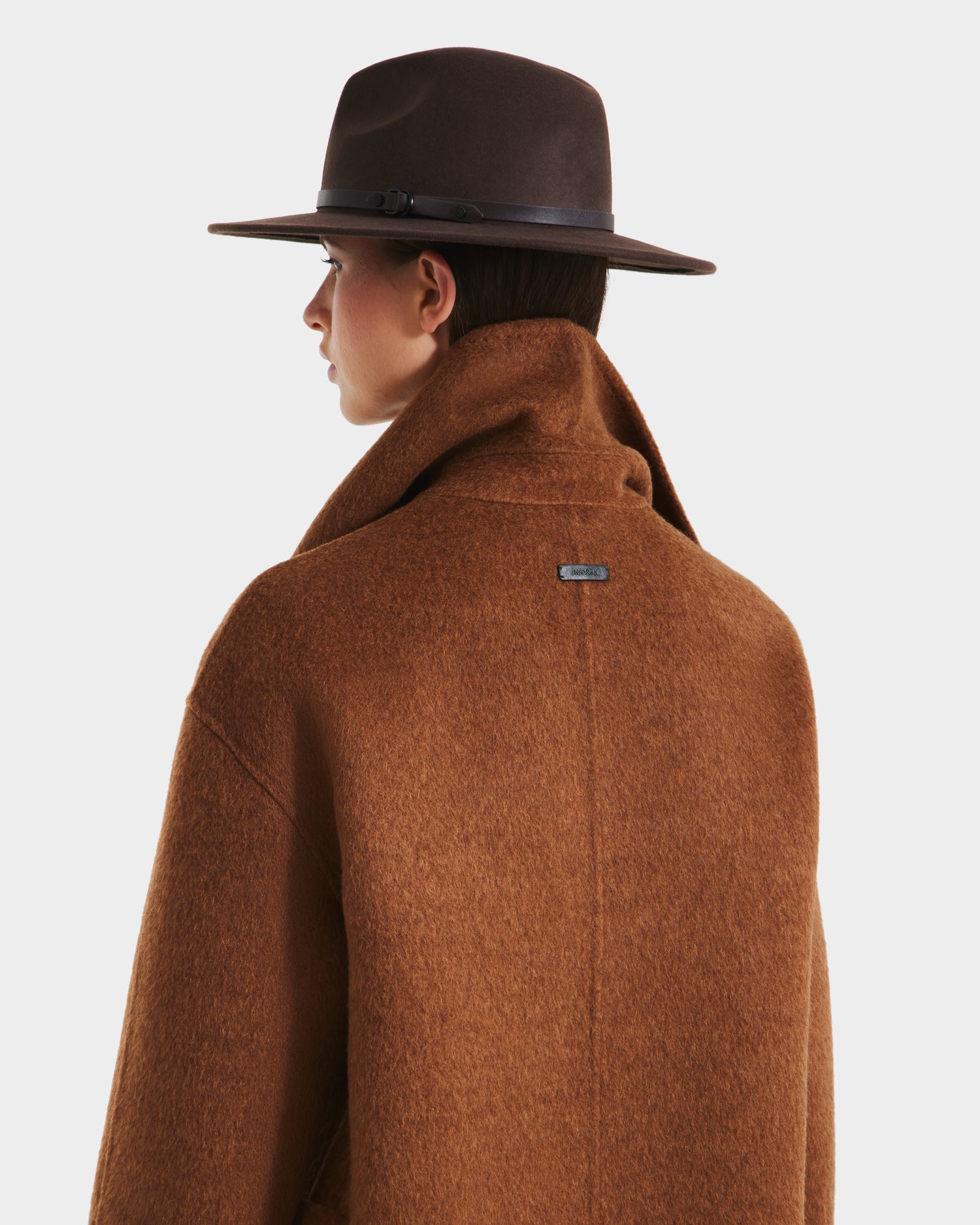 A person in a wide-brimmed brown hat and the Rudsak LUCIA - 8125633 Camel women’s wool jacket, shown from behind and in profile against a plain white background.