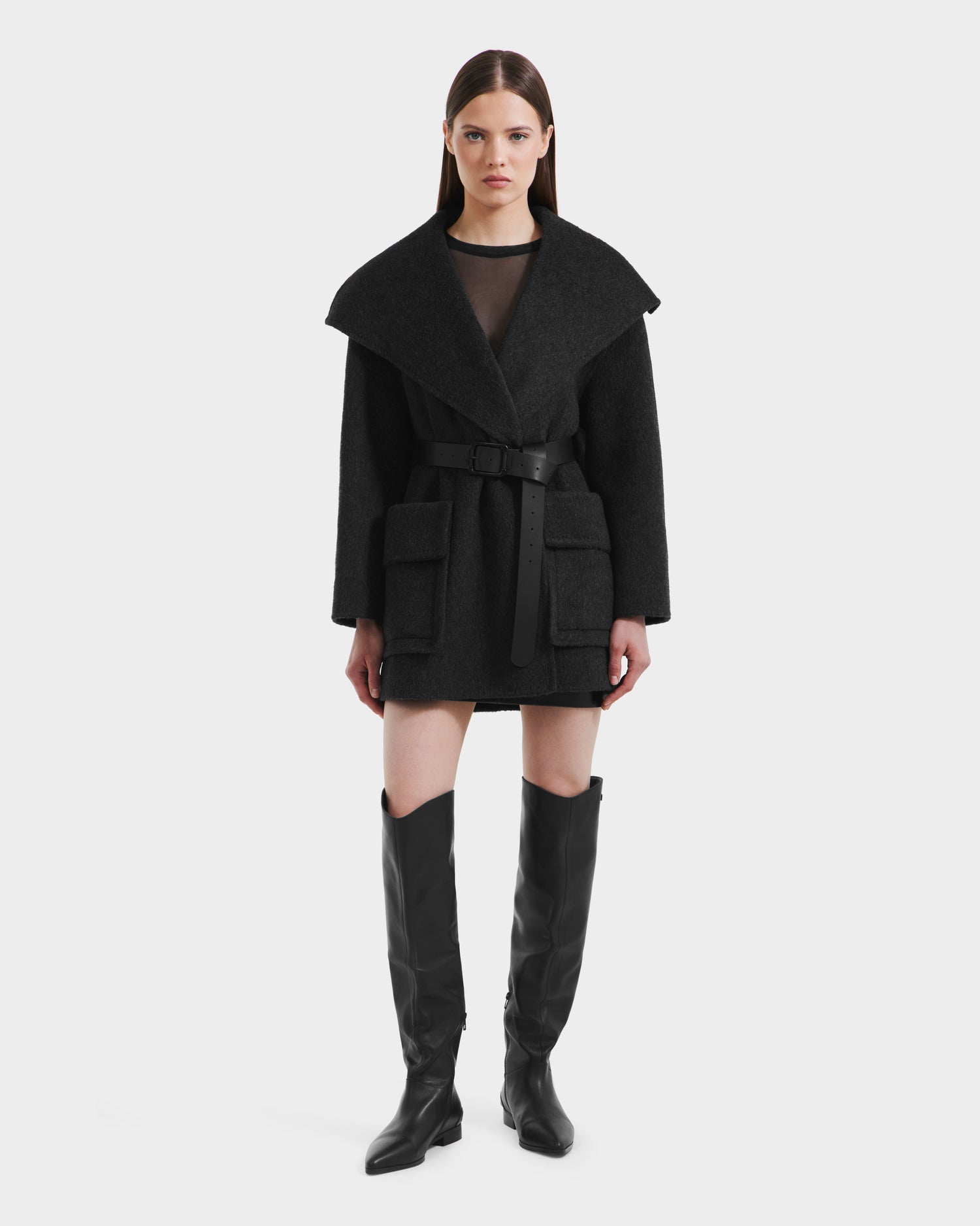 A woman stands facing forward wearing the Rudsak LUCIA - 8125633 Heather Black transitional wool jacket with large lapels, flap pockets, a removable vegan leather belt, and black over-the-knee boots against a plain white background.