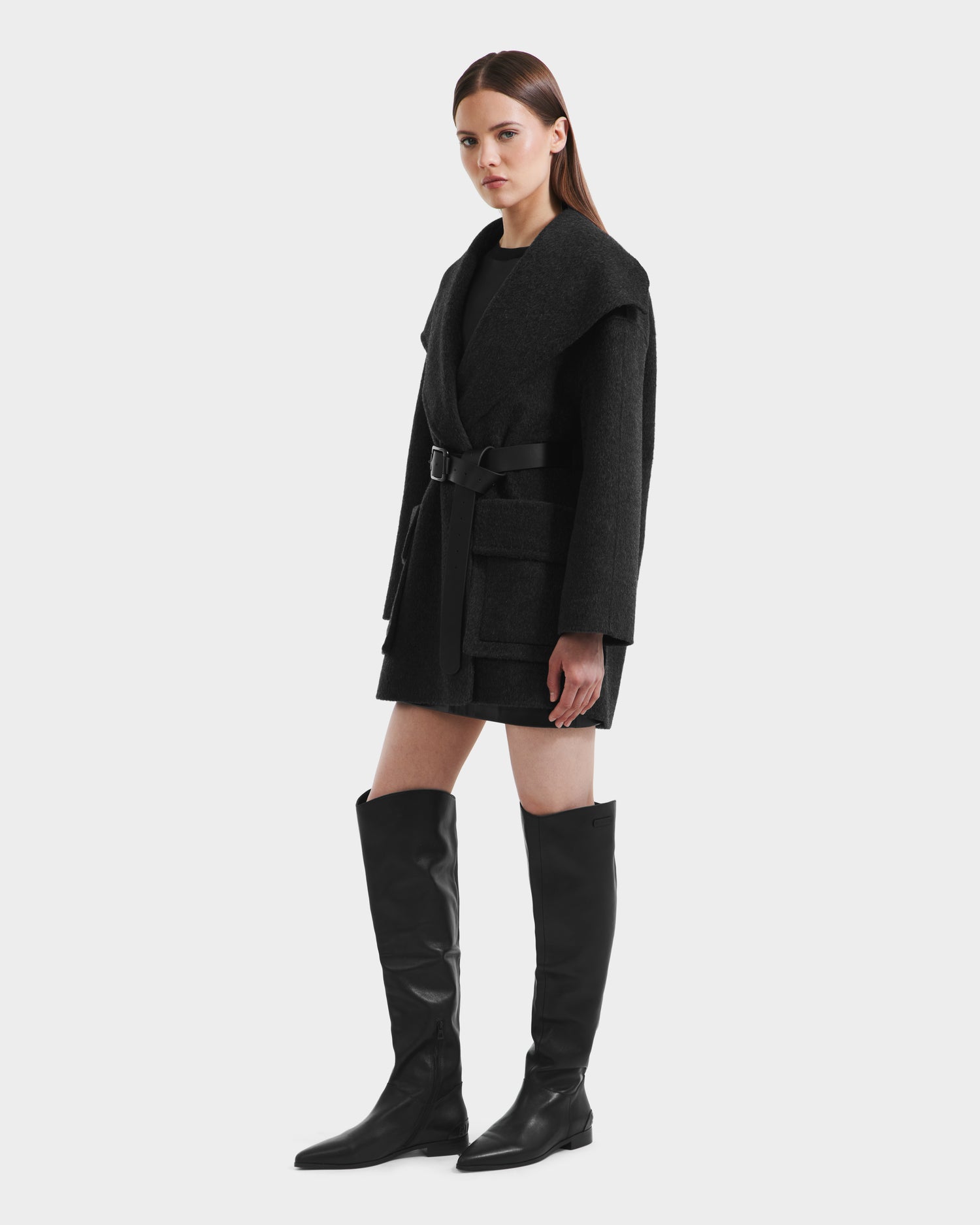 A woman stands facing forward, wearing the Rudsak LUCIA - 8125633 Heather Black transitional wool jacket with large lapels, patch pockets, and a removable vegan leather belt, paired with knee-high black boots against a plain white background.