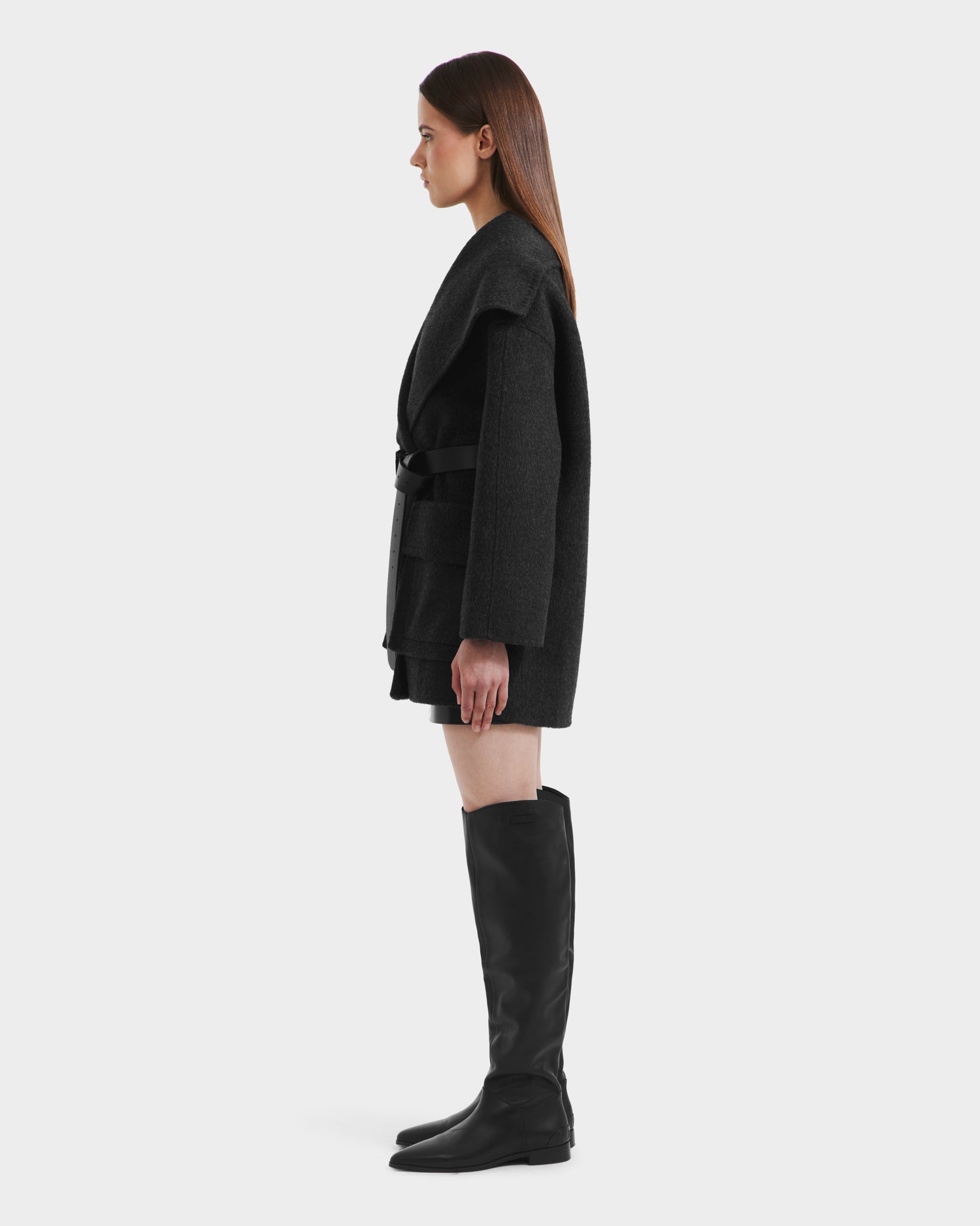 A woman in profile against a plain background wears the Rudsak LUCIA - 8125633 Heather Black women’s transitional wool jacket with a removable vegan leather belt, styled with a short black skirt and tall black boots. Her straight hair falls past her shoulders.