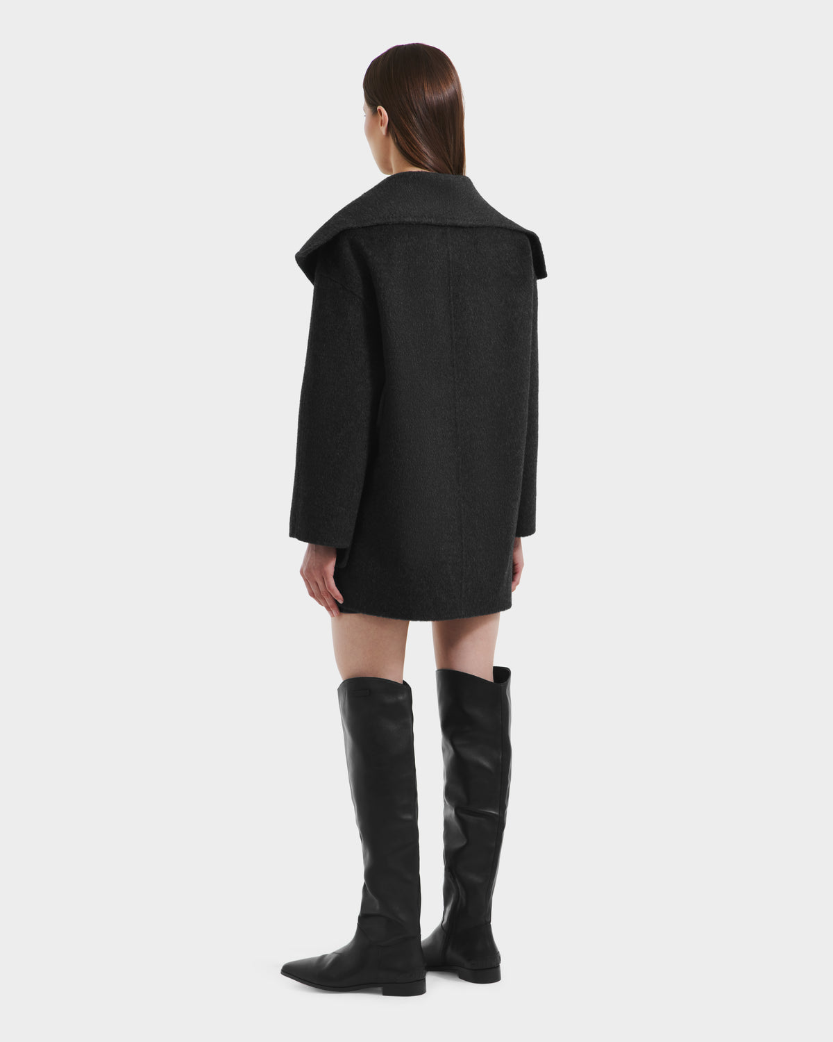 A woman stands facing away in the Rudsak LUCIA - 8125633 Heather Black women’s wool jacket with a wide collar and long sleeves, paired with knee-high black boots. Her straight brown hair is down against a plain white background.