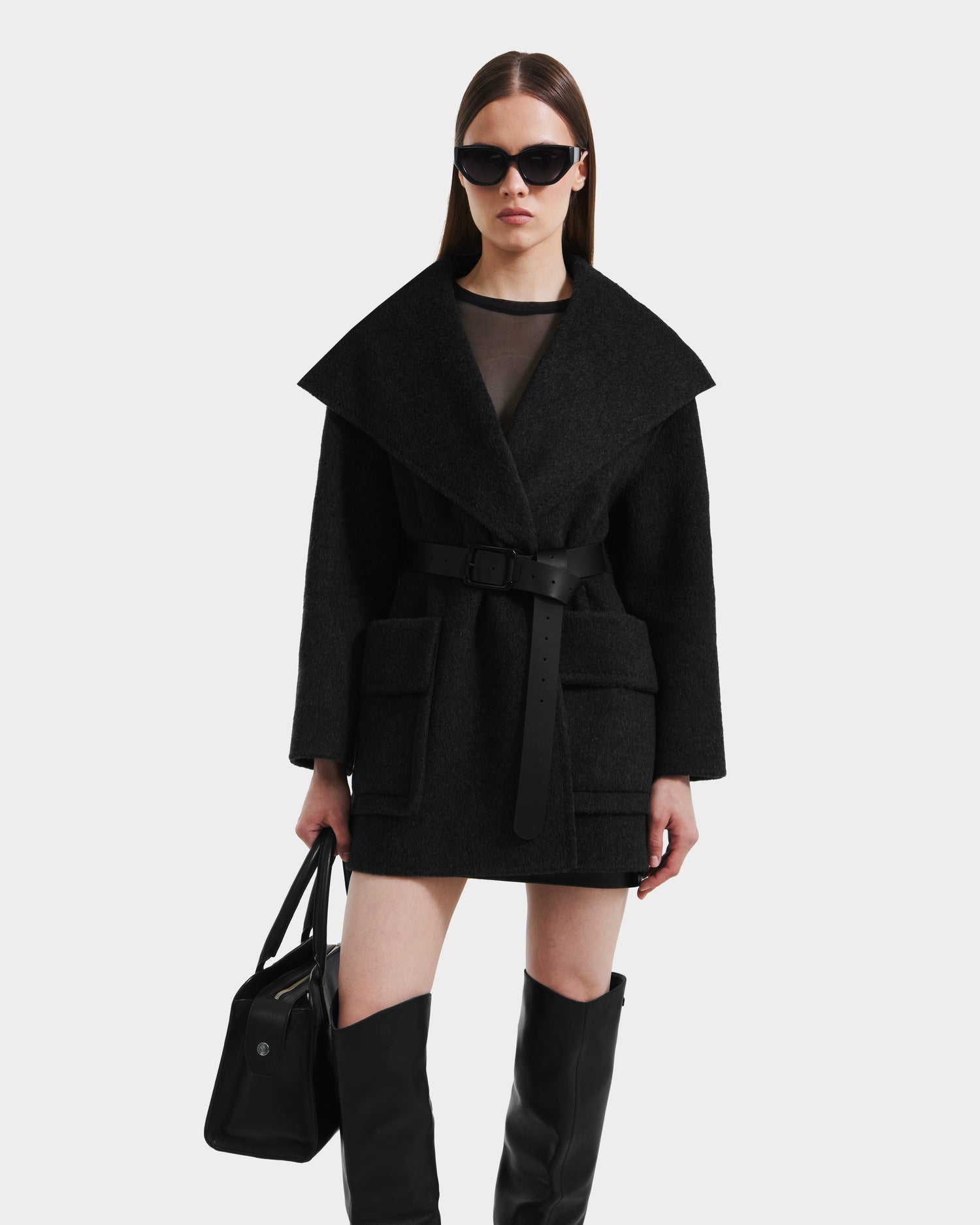 A woman wearing the Rudsak LUCIA - 8125633 Heather Black belted wool coat, styled with black knee-high boots, sunglasses, and a black handbag, stands against a plain white background.