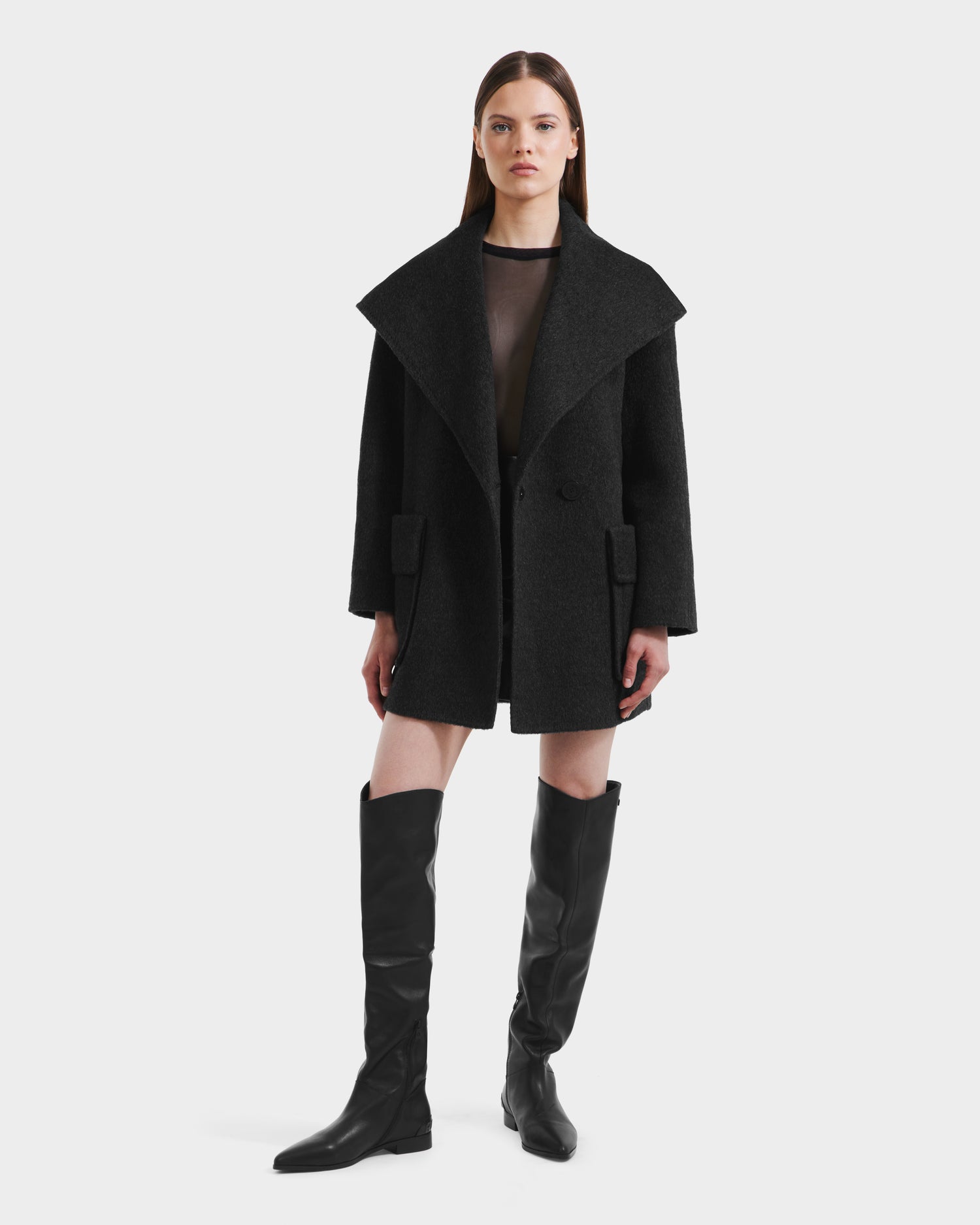 A woman with straight, long brown hair and a neutral expression stands against a white background, wearing the Rudsak LUCIA - 8125633 Heather Black oversized coat with wide lapels and a removable vegan leather belt.