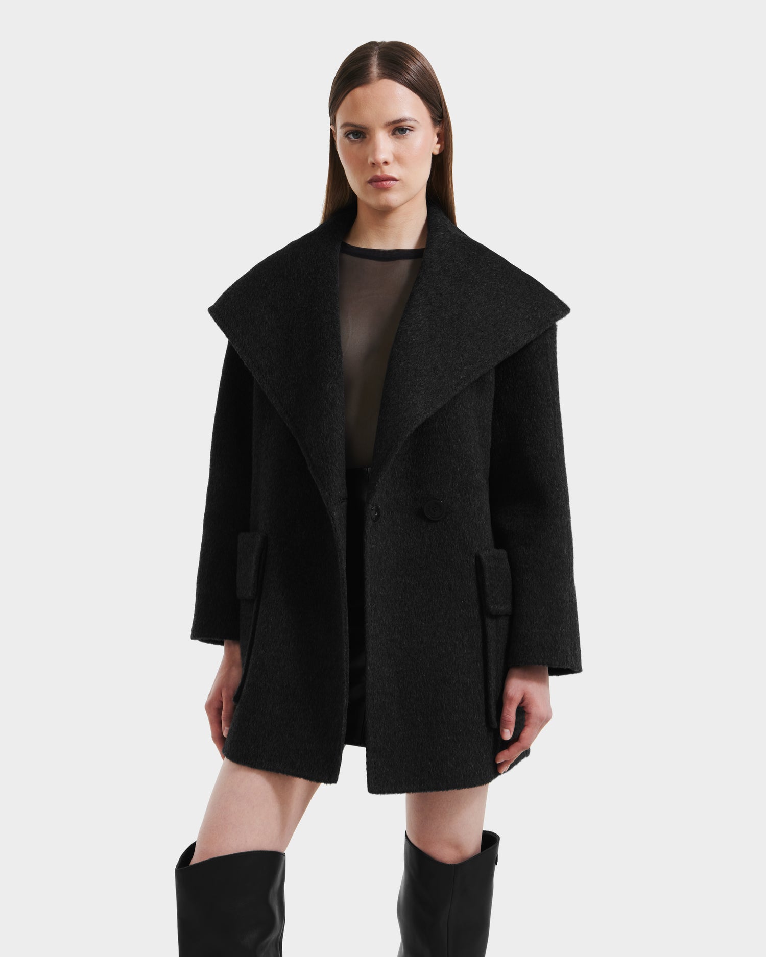 A woman stands against a white background wearing the Rudsak LUCIA - 8125633 Heather Black oversized coat with wide lapels and a removable vegan leather belt, styled with a sheer black top and black knee-high boots.