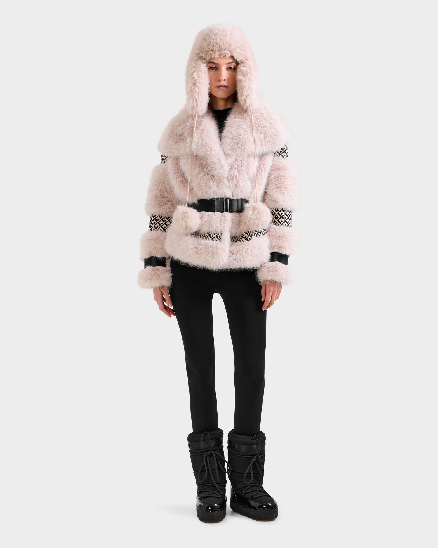 A person models the Rudsak LUELLA - 8125656 Vanilla Monogram faux fur coat with hood and black belt, paired with black leggings and winter boots—an eye-catching winter statement piece against a plain light background.