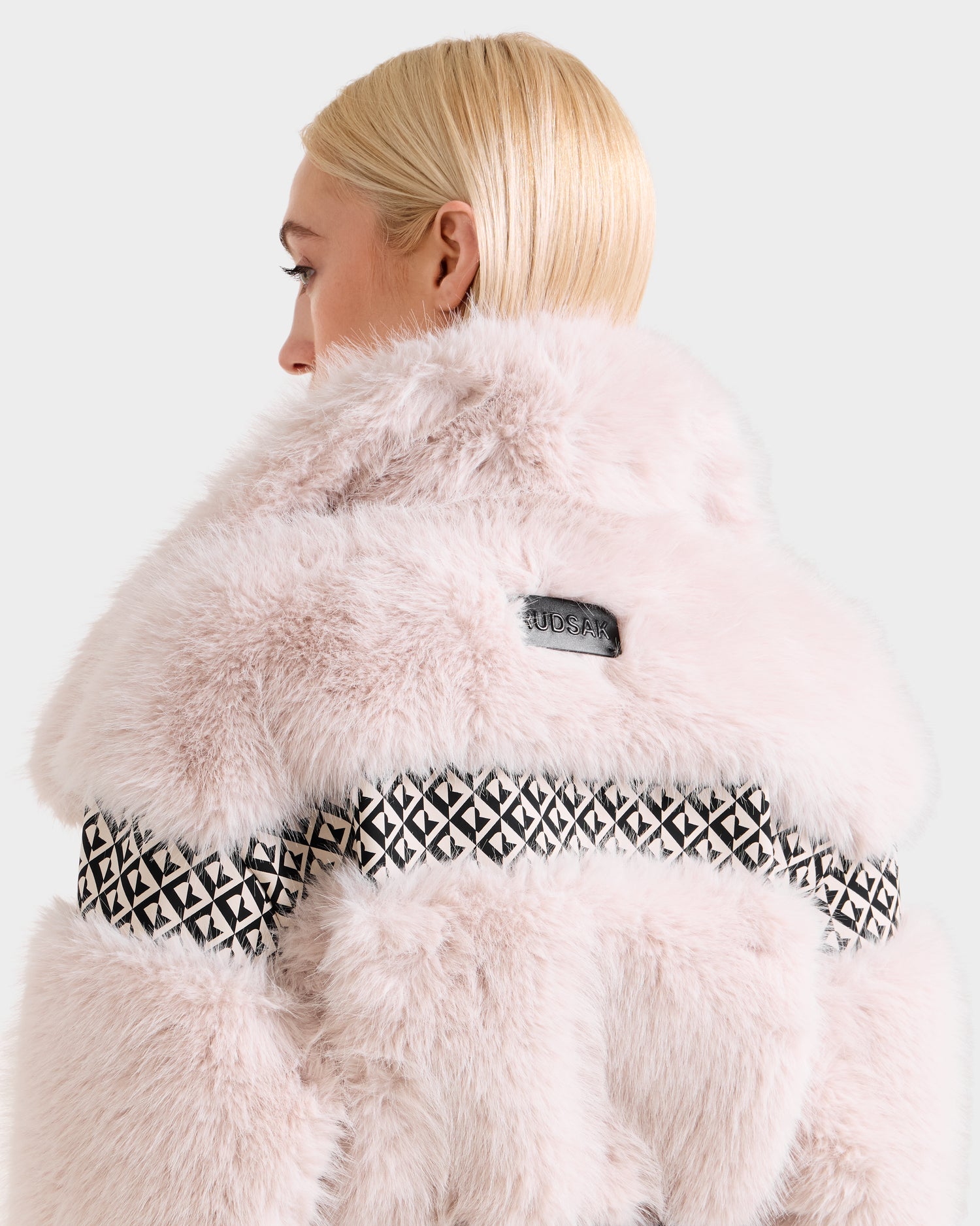 A woman with straight blonde hair, seen from behind, wears the Rudsak LUELLA - 8125656 Vanilla Monogram faux fur coat featuring a black geometric band—a striking and stylish winter statement.