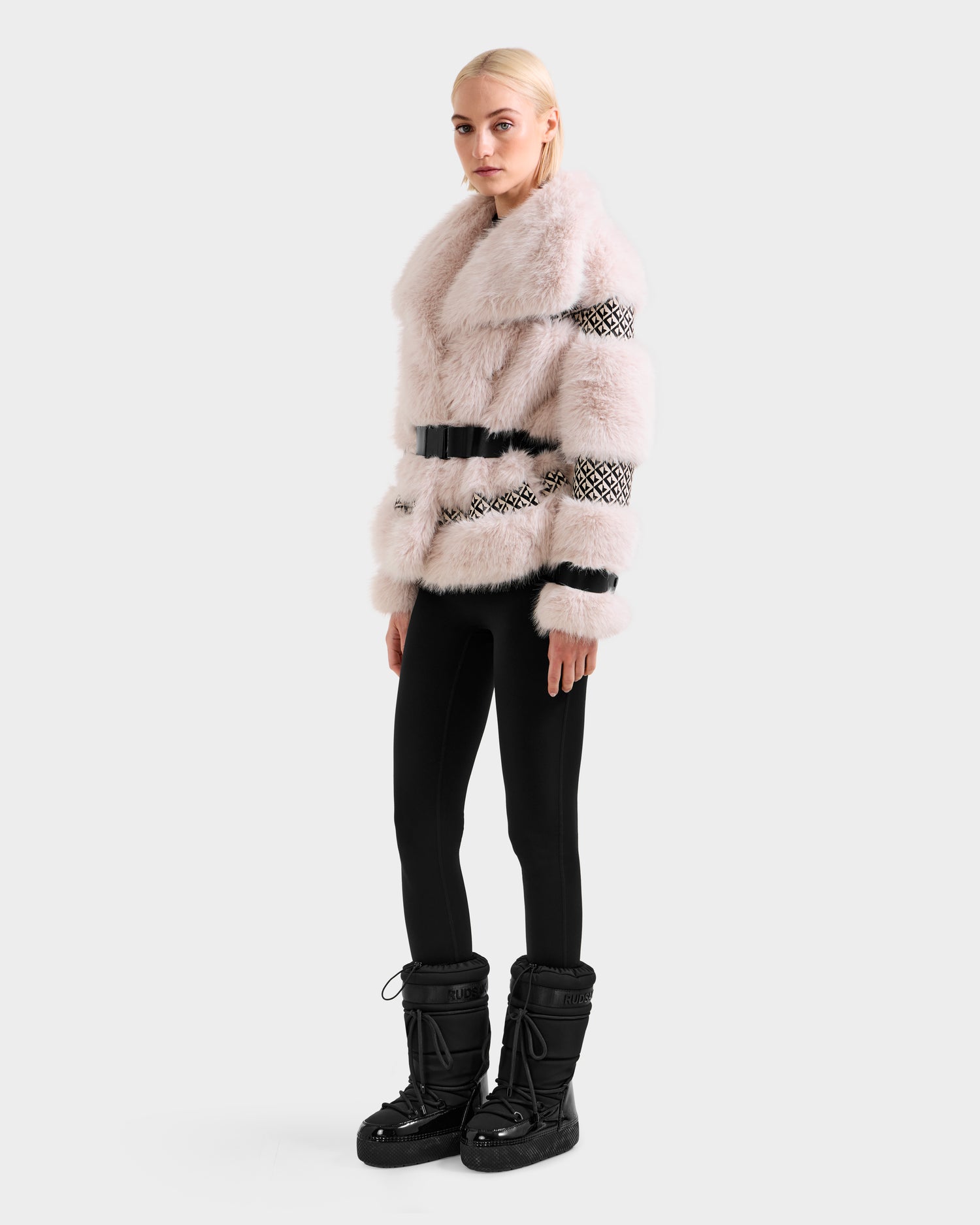 A woman with blonde hair wears the Rudsak LUELLA - 8125656 Vanilla Monogram faux fur coat, styled with a black belt, black leggings, and winter boots, standing against a plain light background.