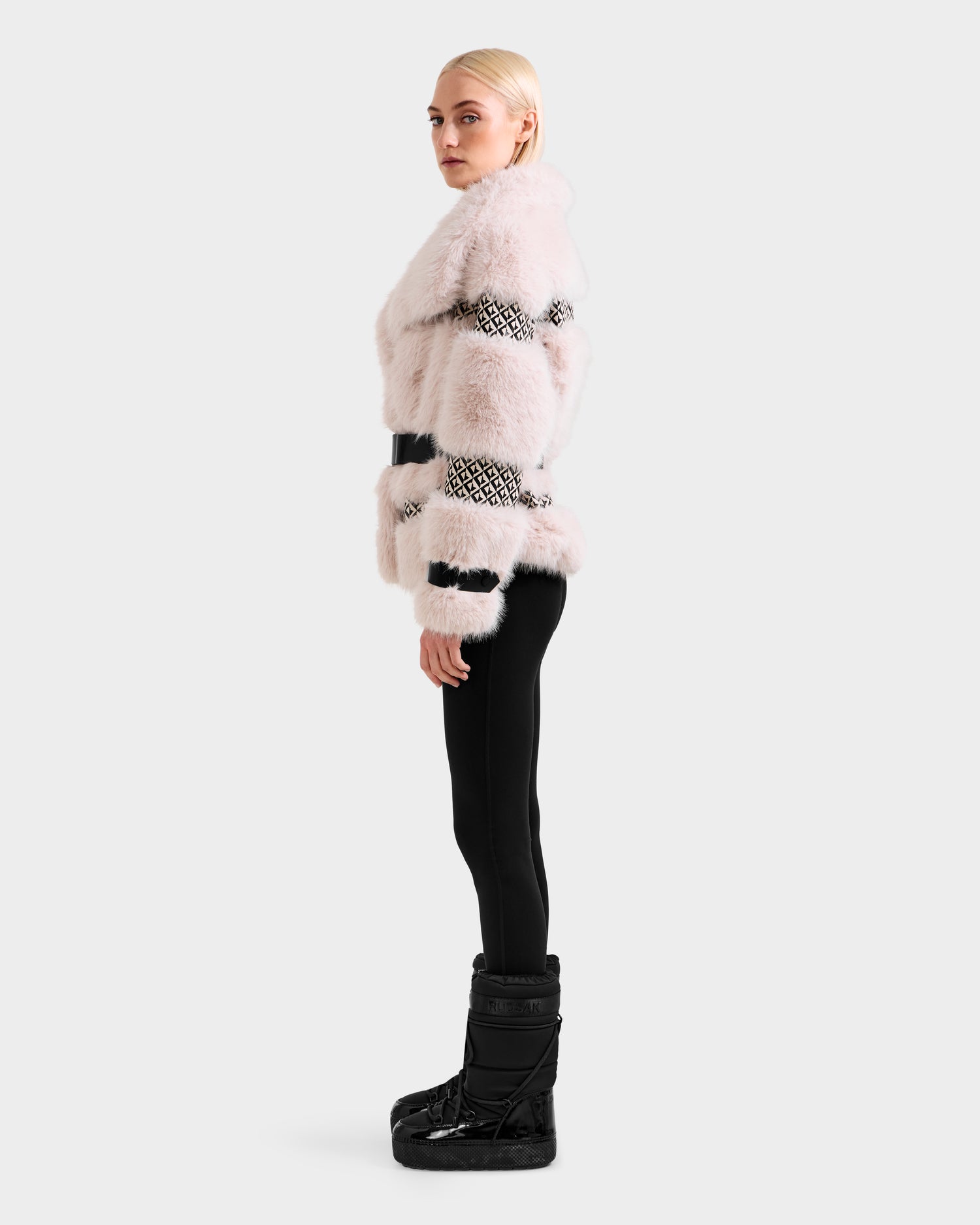 A woman with blonde hair stands in profile wearing the Rudsak LUELLA - 8125656 Vanilla Monogram coat, paired with black leggings and boots, against a plain light gray background.