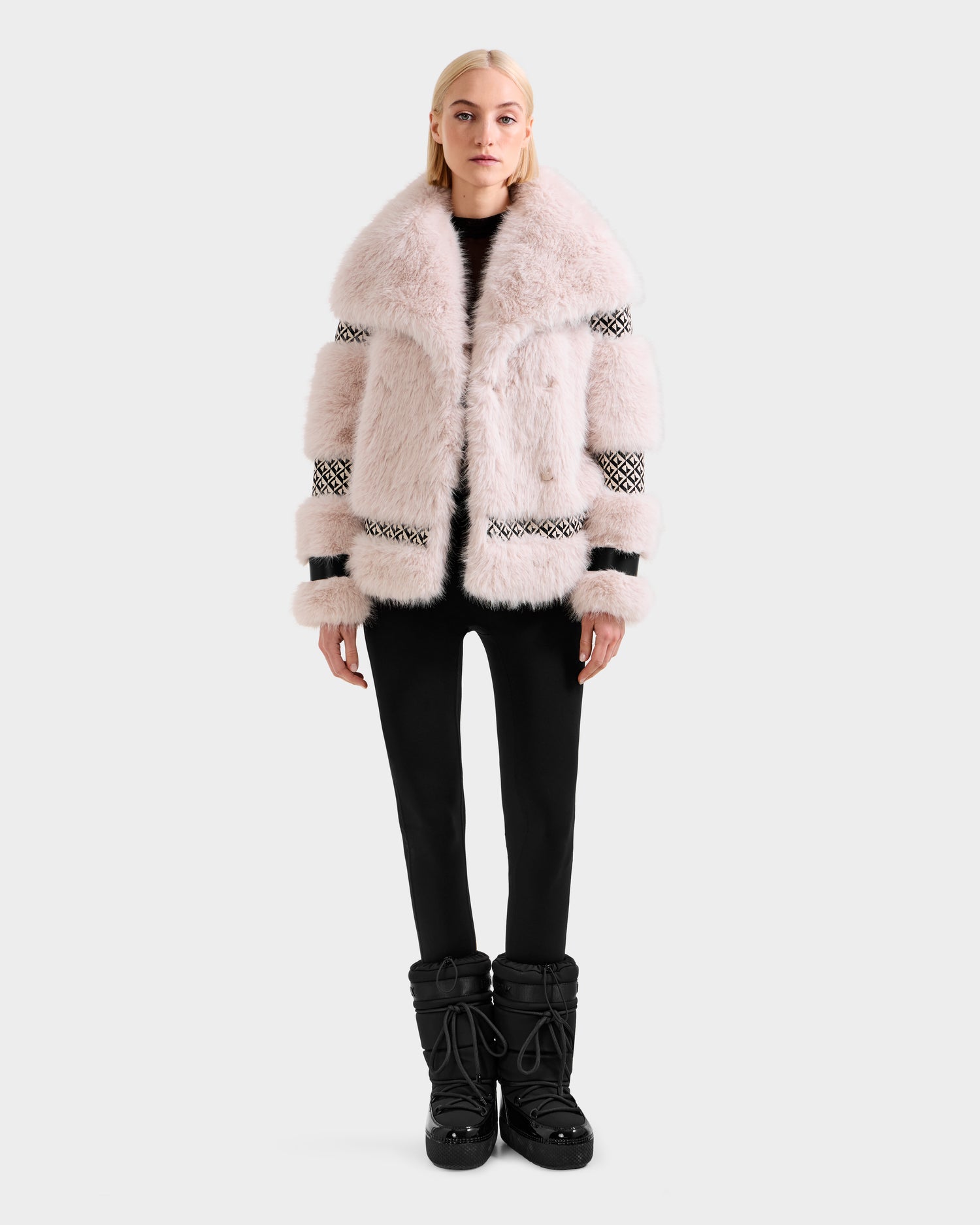 A woman faces forward in the Rudsak LUELLA - 8125656 Vanilla Monogram coat, featuring black and white patterned bands, paired with black pants and winter boots against a light gray backdrop for a chic winter statement look.