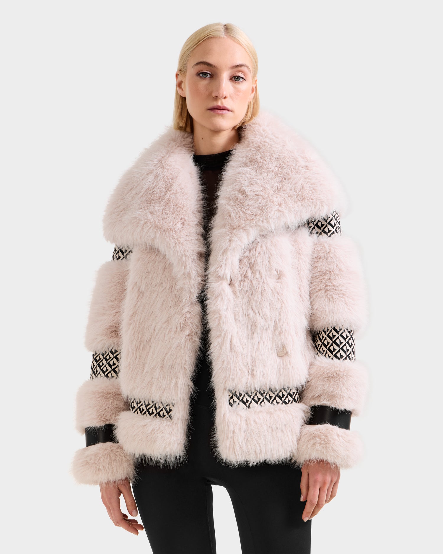 A woman with straight blonde hair models the Rudsak LUELLA - 8125656 Vanilla Monogram light pink faux fur coat with black and white sleeve and body accents over a black outfit, standing against a plain light background.