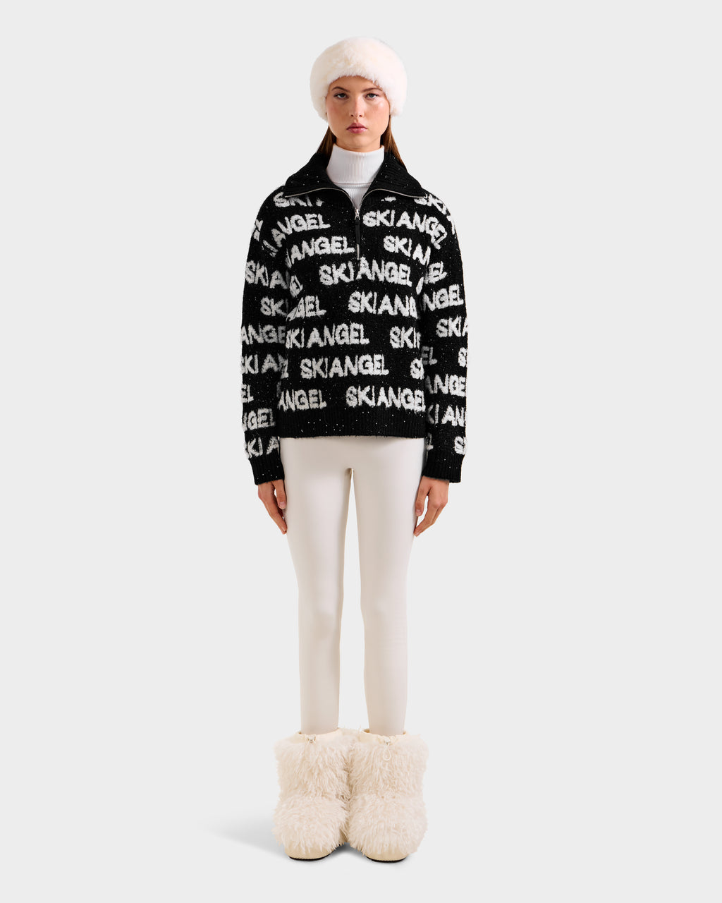 A woman stands against a light gray background wearing Rudsak’s LYDIA - 8525687 Black, a stylish women’s pullover, paired with cream leggings, a white fur hat, and fluffy boots.