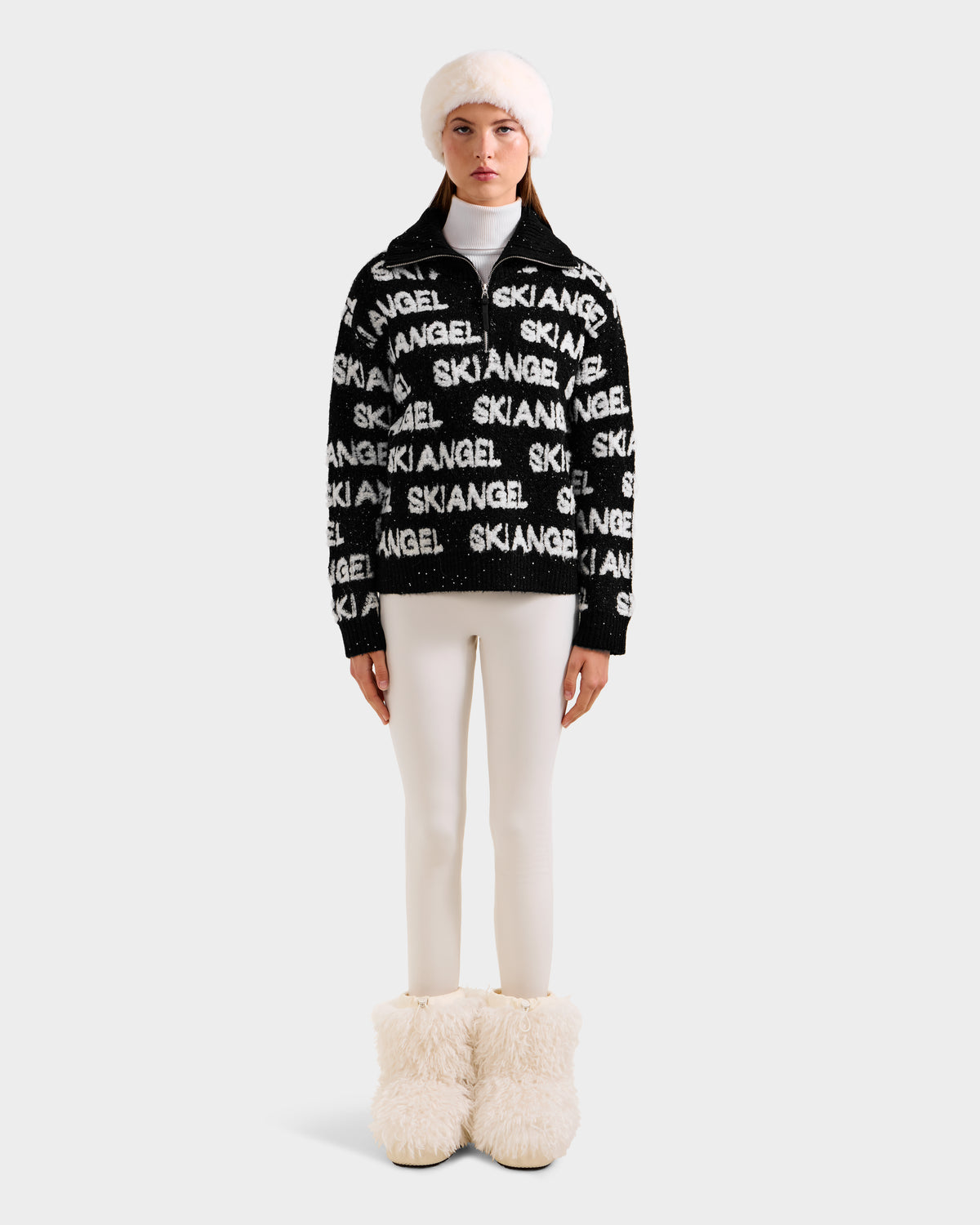 A woman stands against a light gray background wearing Rudsak’s LYDIA - 8525687 Black, a stylish women’s pullover, paired with cream leggings, a white fur hat, and fluffy boots.