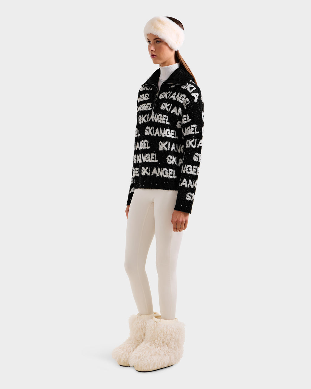 A woman wears the Rudsak LYDIA - 8525687 Black sweater with white leggings, fluffy white boots, and a white fur headband, standing against a plain light background.