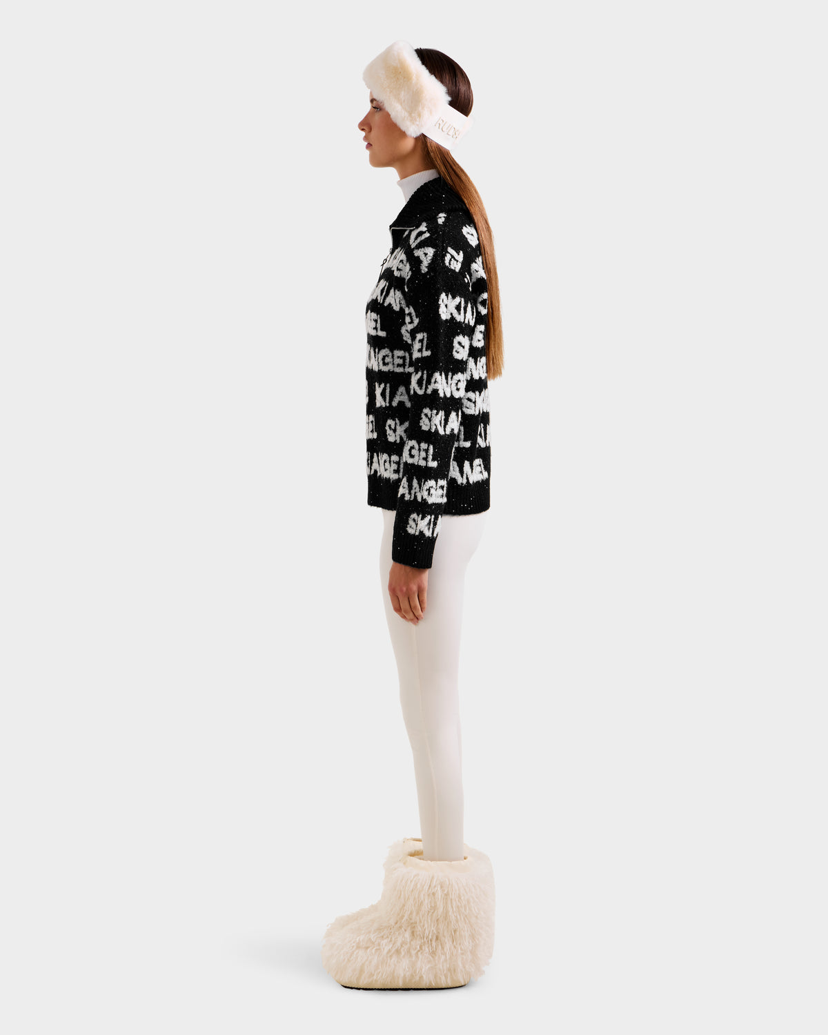 A woman in profile wears a black Rudsak LYDIA - 8525687 sweater with white SKI ANGEL text, paired with white leggings, fluffy earmuffs, and large fuzzy boots against a light background.