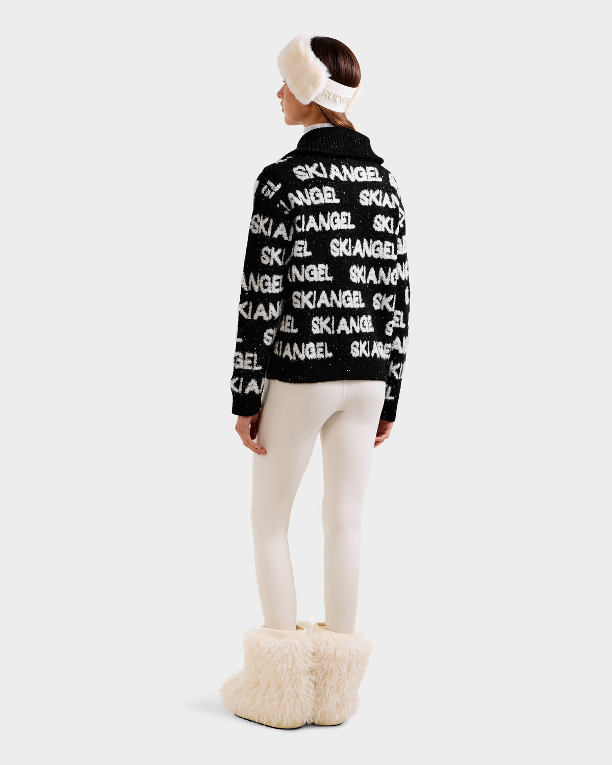 A woman faces away, wearing the Rudsak LYDIA - 8525687 Black sweater with white text, paired with white leggings, fluffy cream boots, and a white fur headband against a plain light background.