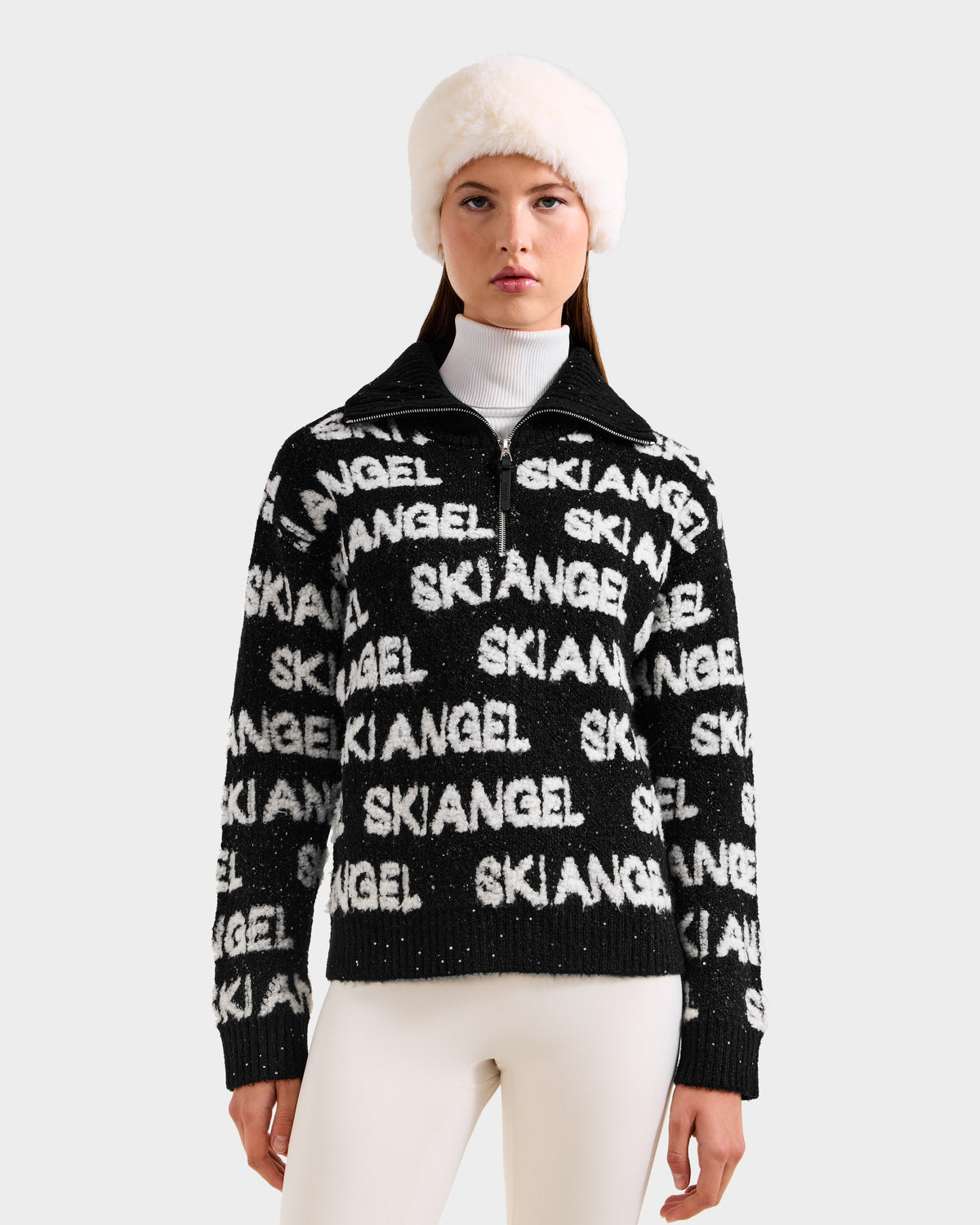A woman models the Rudsak LYDIA - 8525687 Black women’s sweater with “SKI ANGEL” in white, styled with a turtleneck, white pants, and a fluffy hat against a light gray background—ideal for a chic winter wardrobe.
