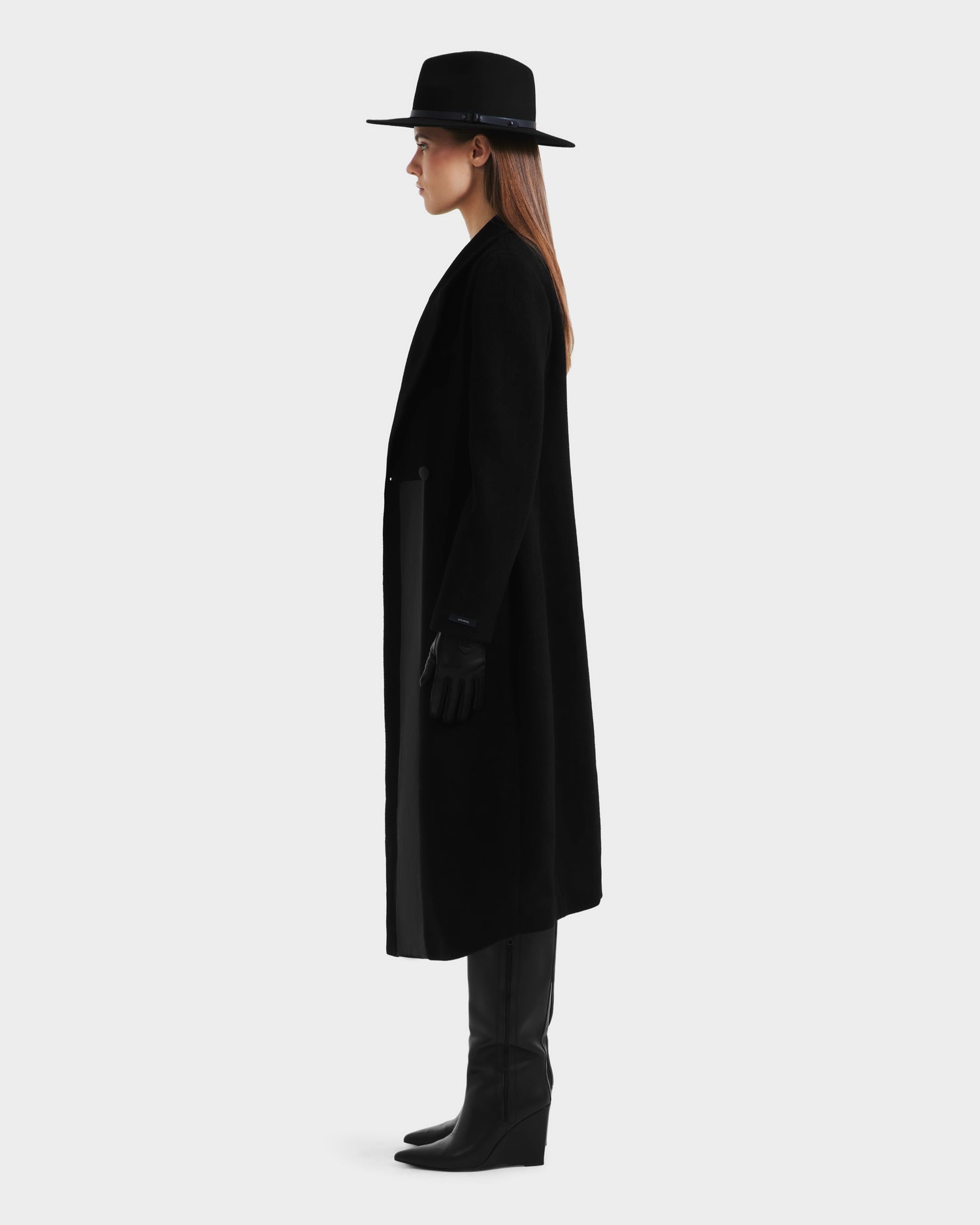 A woman stands in profile wearing the LYNWOOD - 8125539 Black coat by Rudsak, styled with a black wide-brim hat, black gloves, and knee-high black boots against a plain light background.