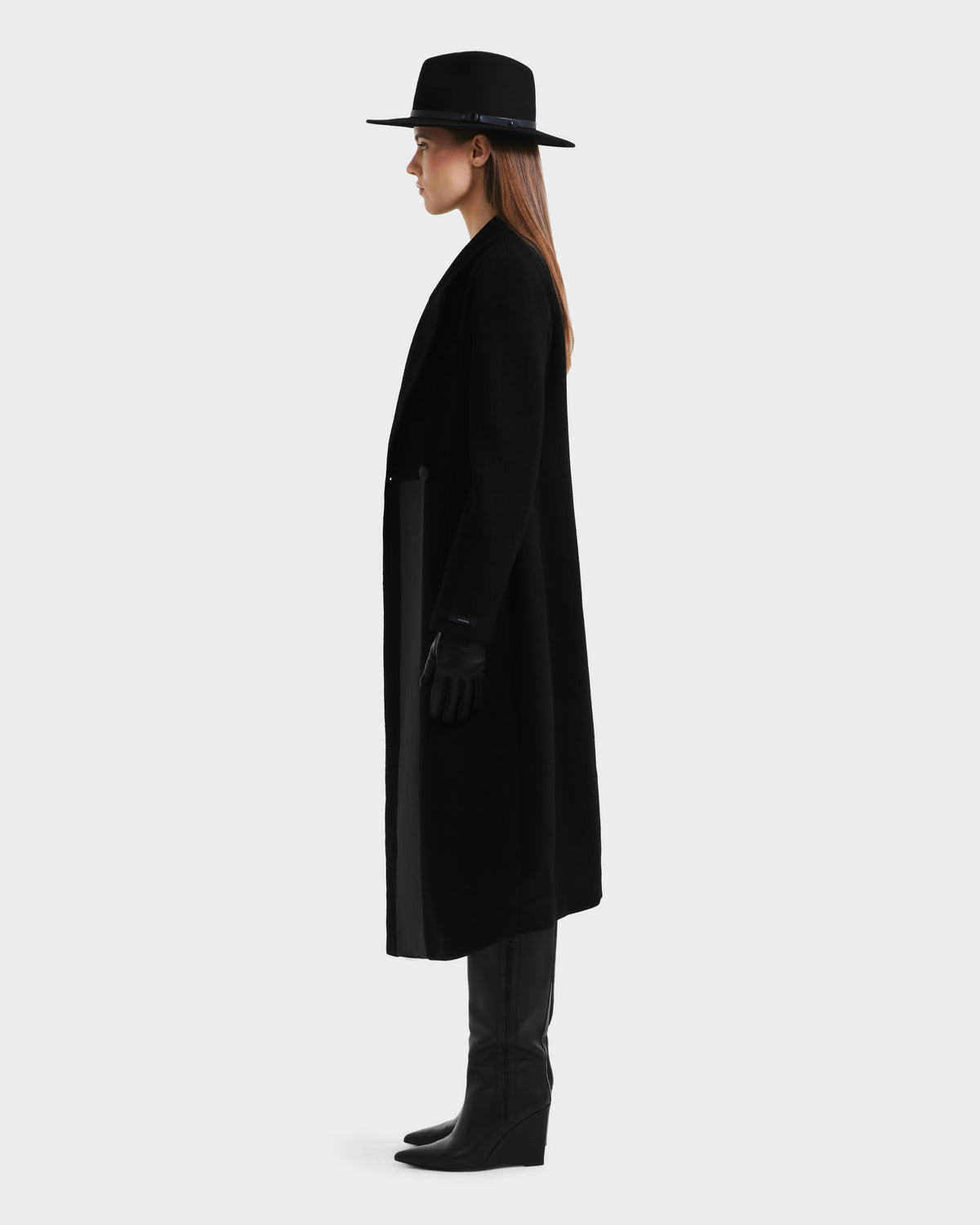 A woman stands in profile wearing the LYNWOOD - 8125539 Black coat by Rudsak, styled with a black wide-brim hat, black gloves, and knee-high black boots against a plain light background.