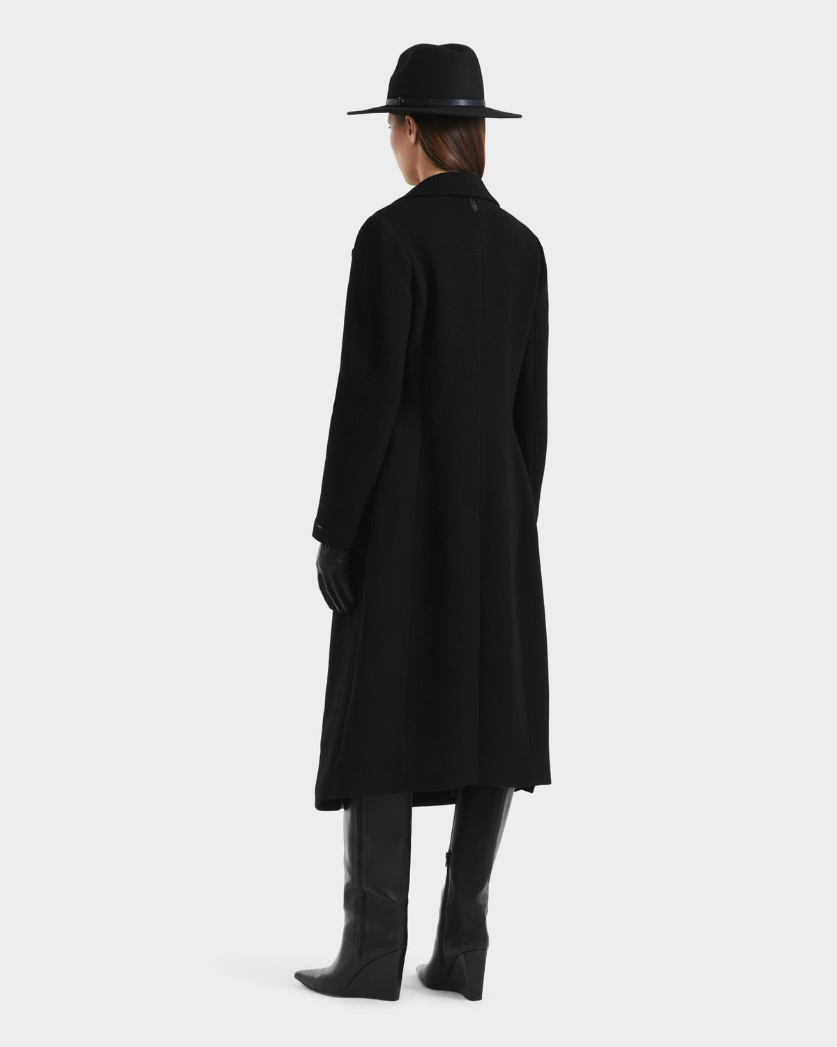 Wearing the Rudsak LYNWOOD - 8125539 Black coat, black gloves, knee-high boots, and a wide-brimmed hat, a person stands facing away against a plain light background.