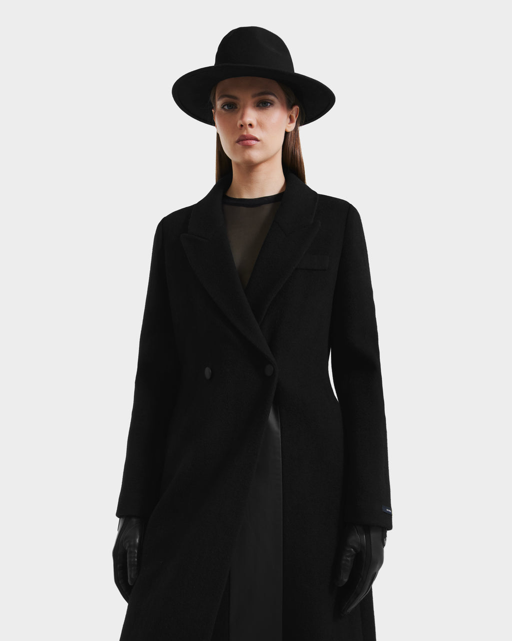 A woman in a black wide-brimmed hat, gloves, and sheer black top stands facing forward against a white background, elegantly styled in the Rudsak LYNWOOD - 8125539 Black womens long tailored coat.