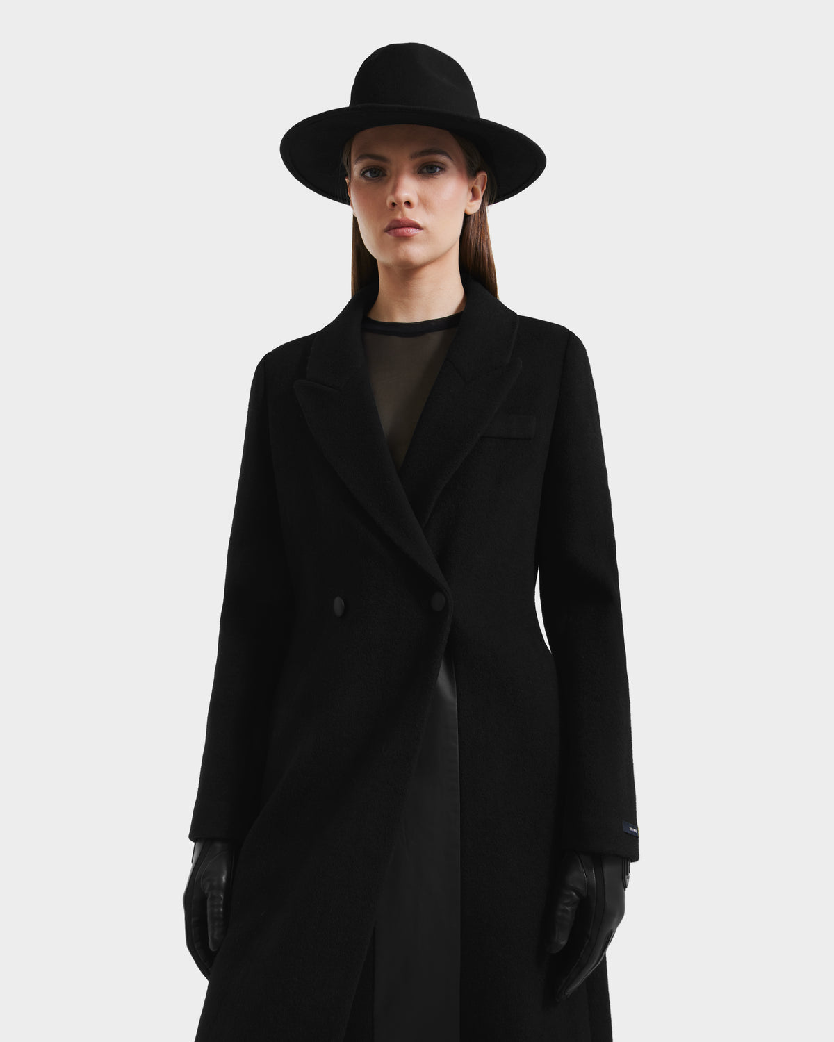 A woman in a black wide-brimmed hat, gloves, and sheer black top stands facing forward against a white background, elegantly styled in the Rudsak LYNWOOD - 8125539 Black womens long tailored coat.