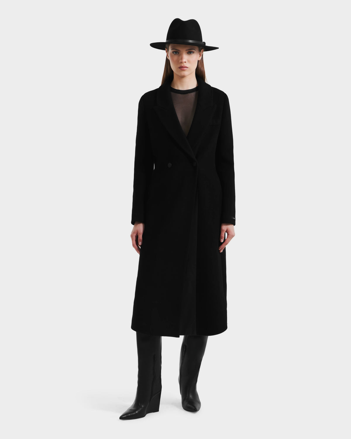 A woman faces forward in a Rudsak LYNWOOD - 8125539 Black long tailored coat, paired with a black wide-brim hat, sheer black top, and knee-high boots against a plain white background.