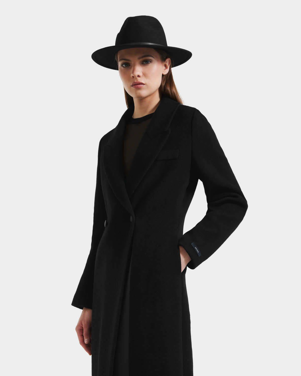 A woman wearing the Rudsak LYNWOOD - 8125539 Black wool coat and a wide-brimmed black hat stands against a white background with one hand in her pocket and a neutral expression.
