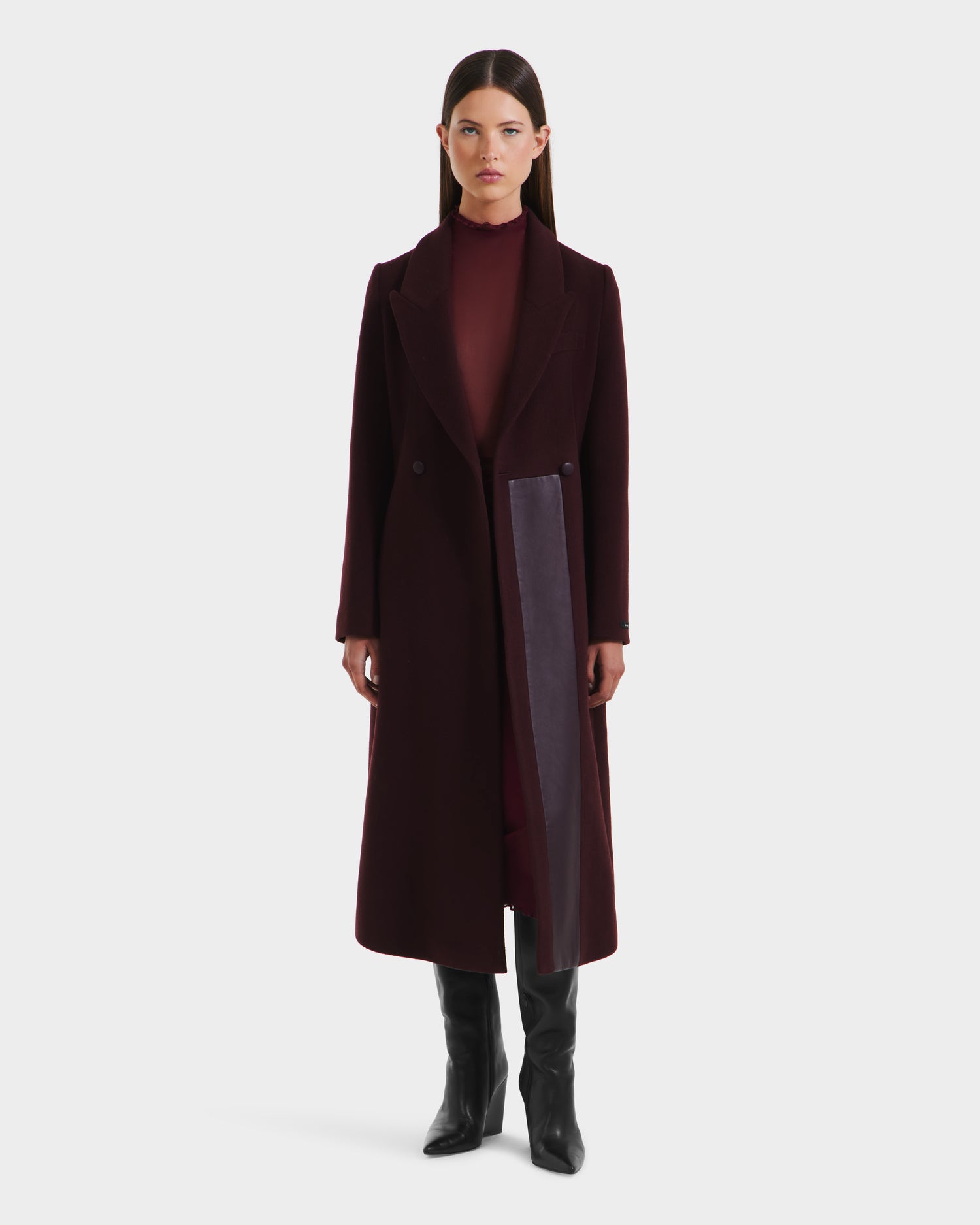 A woman stands facing forward wearing the Rudsak LYNWOOD - 8125539 Bordeaux, a womens long tailored coat in dark burgundy with a leather front panel, over a matching outfit and black knee-high boots, set against a plain white background.