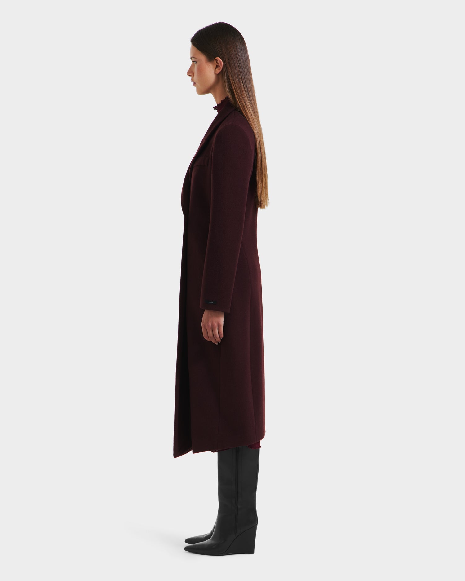 A woman in profile faces left, wearing the Rudsak LYNWOOD - 8125539 Bordeaux long tailored coat with black knee-high boots and straight brown hair, set against a plain white background.